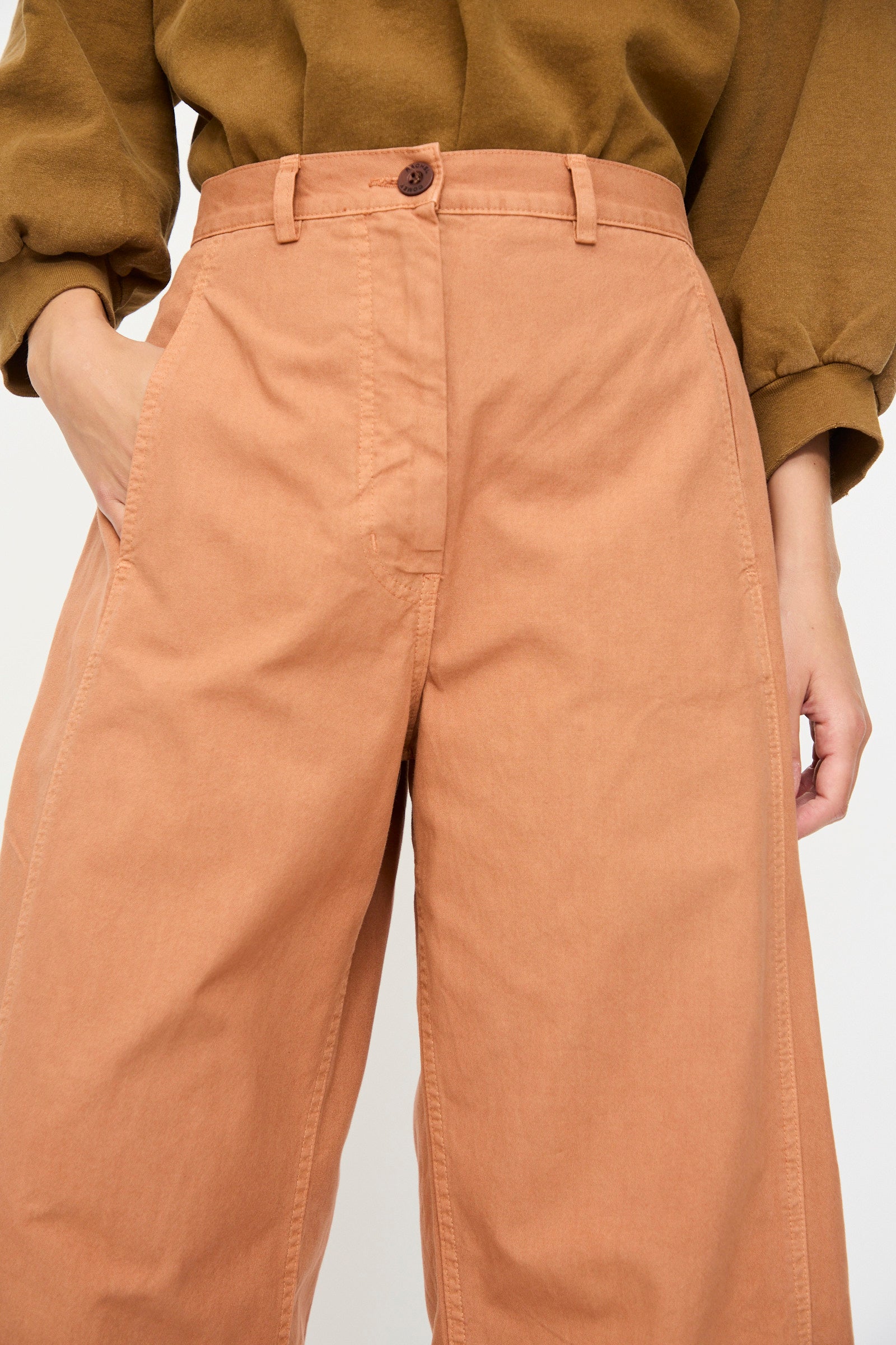 Model wearing the Rachel Comey Garra Pant in Copper, styled as high-waisted wide-leg pants, paired with an olive green long-sleeve top, one hand in pocket; visible from waist to mid-thigh.