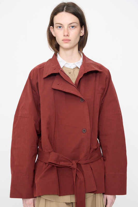 A person wears the Rachel Comey Gosland Trench in Garnet, a rust-colored water-repellent relaxed-fit jacket, over a beige collared shirt while standing against a plain white background.