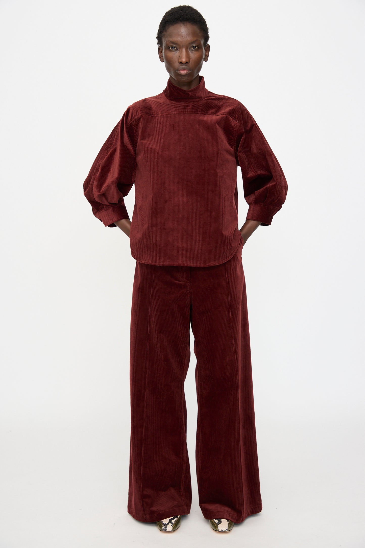 A person stands facing forward in the Rachel Comey Groth Top in Brick, a loose-fitting, high mock neck velvet top with quarter sleeves, paired with matching wide-leg pants, hands in pockets against a plain white background.
