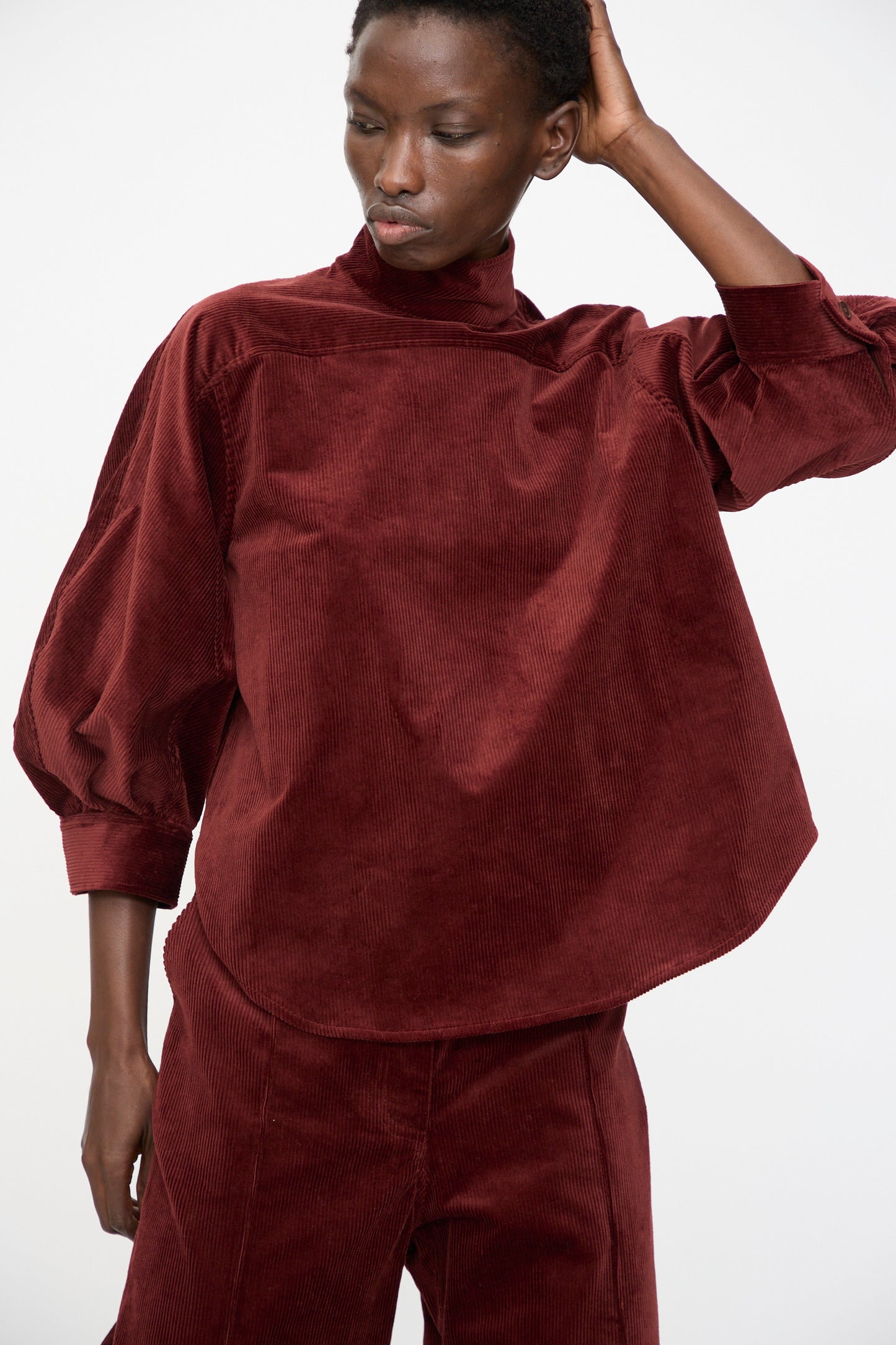 A person wears the Rachel Comey Groth Top in Brick, a loose-fitting stretch corduroy style with a high mock neck, and stands with one hand behind their head against a plain background.
