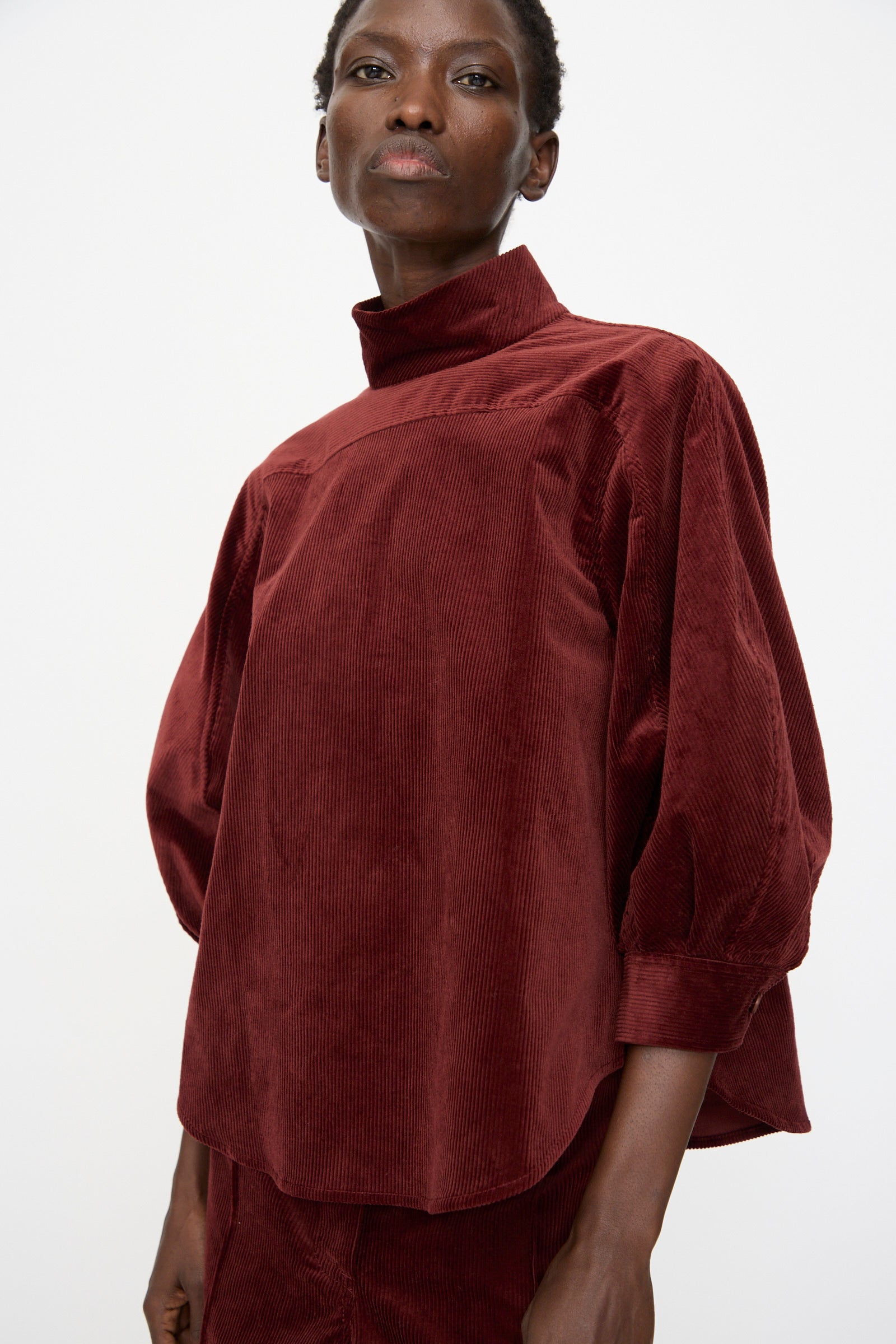 A person wears the Rachel Comey Groth Top in Brick, a loose-fitting, stretch corduroy style with a high mock neck and long sleeves, standing against a plain white background.