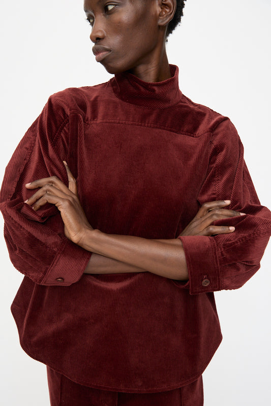 Model wearing the Rachel Comey Groth Top in Brick, featuring a high mock neck and long sleeves, stands with arms crossed and looks to the side against a plain background.