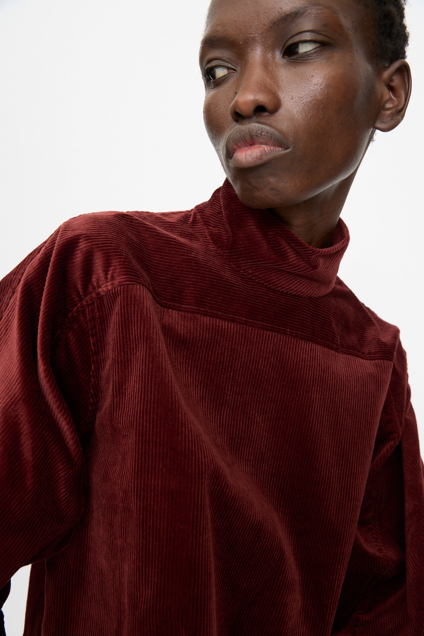 A person wears the Rachel Comey Groth Top in Brick, a long-sleeved, high mock neck stretch corduroy piece, looking to the side against a plain background.