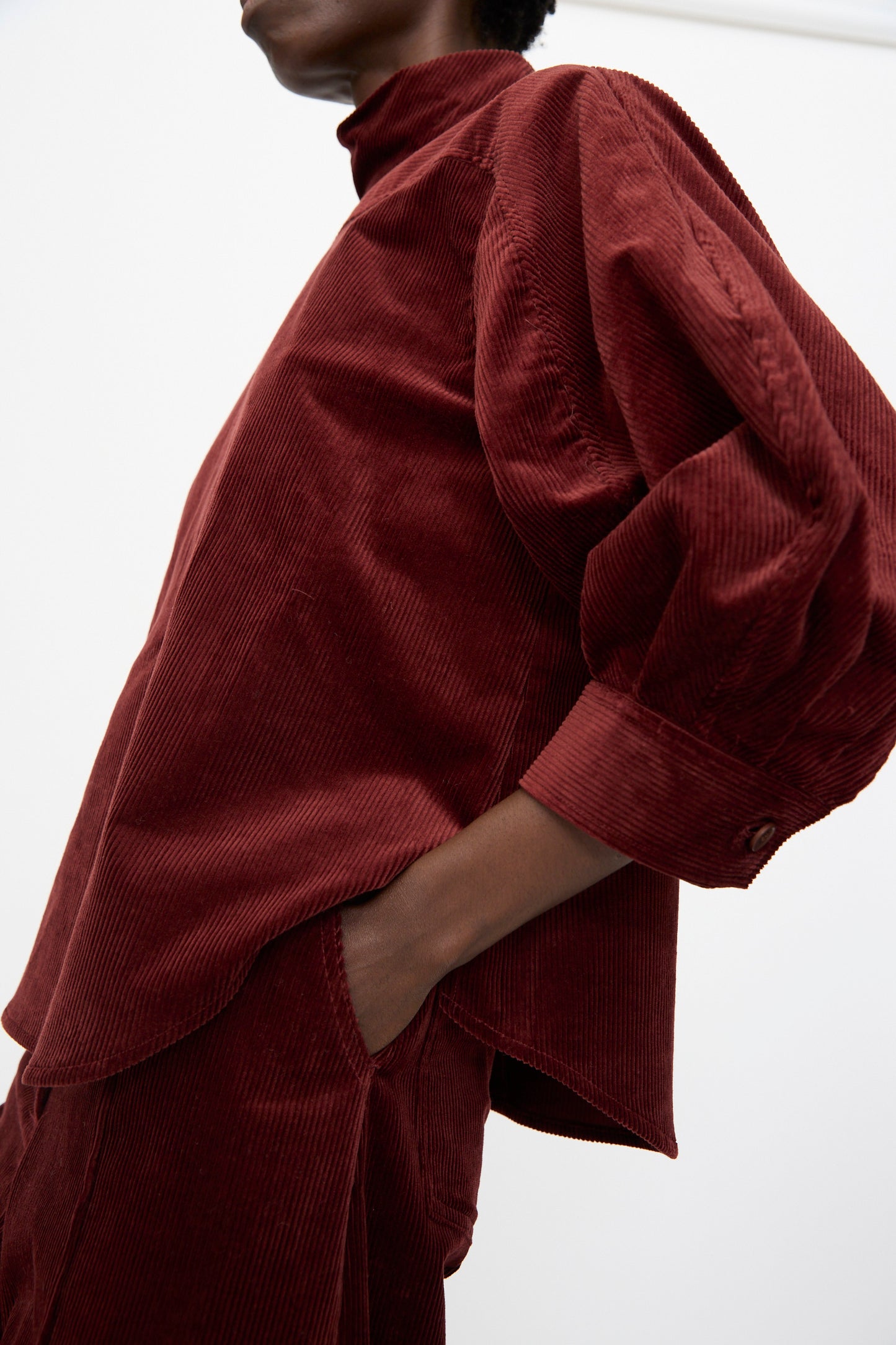 A person wearing the Rachel Comey Groth Top in Brick, a dark red stretch corduroy with puffed quarter-sleeves, paired with matching pants, stands with one hand in their pocket against a plain background.
