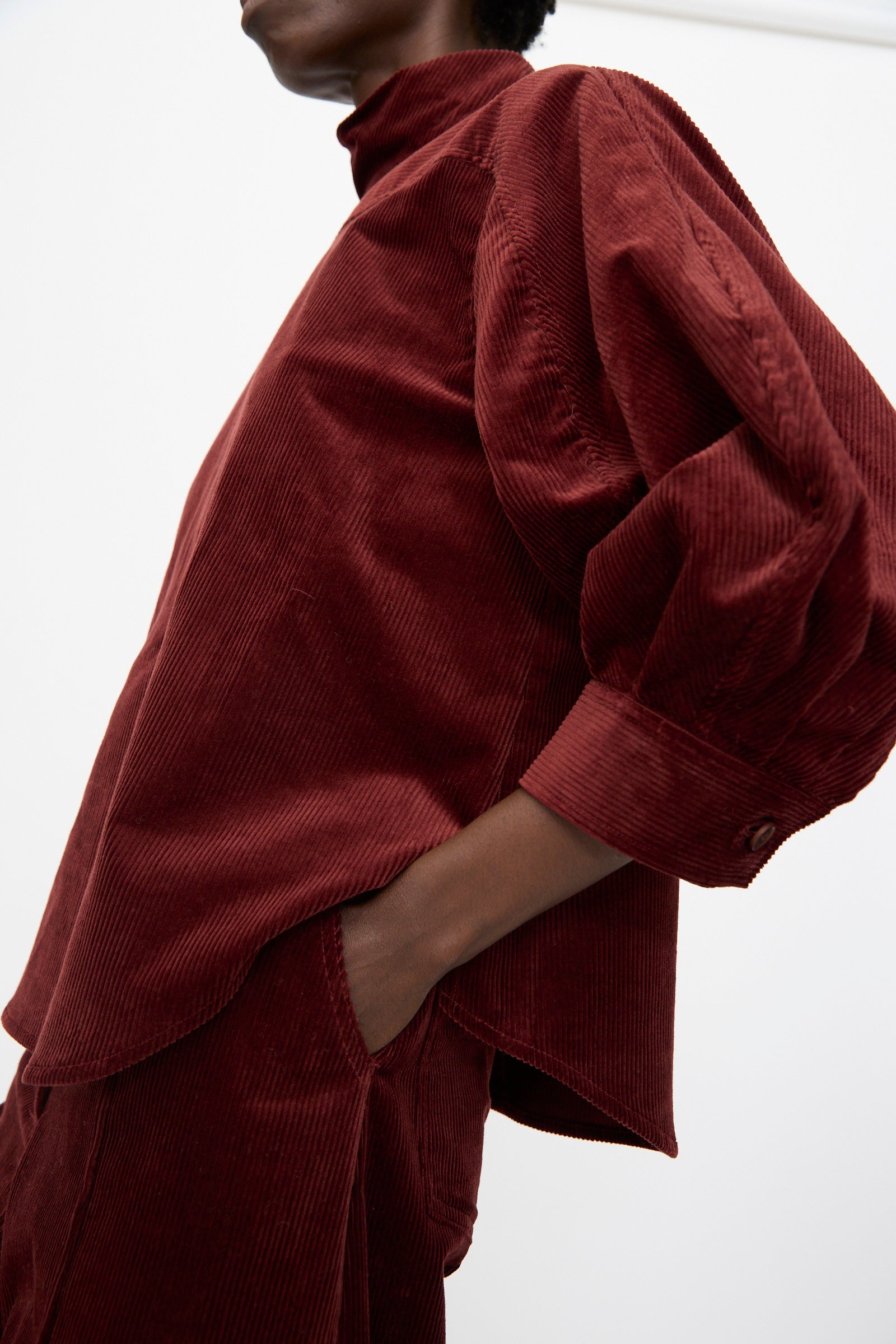A person wearing the Rachel Comey Groth Top in Brick, a dark red stretch corduroy with puffed quarter-sleeves, paired with matching pants, stands with one hand in their pocket against a plain background.