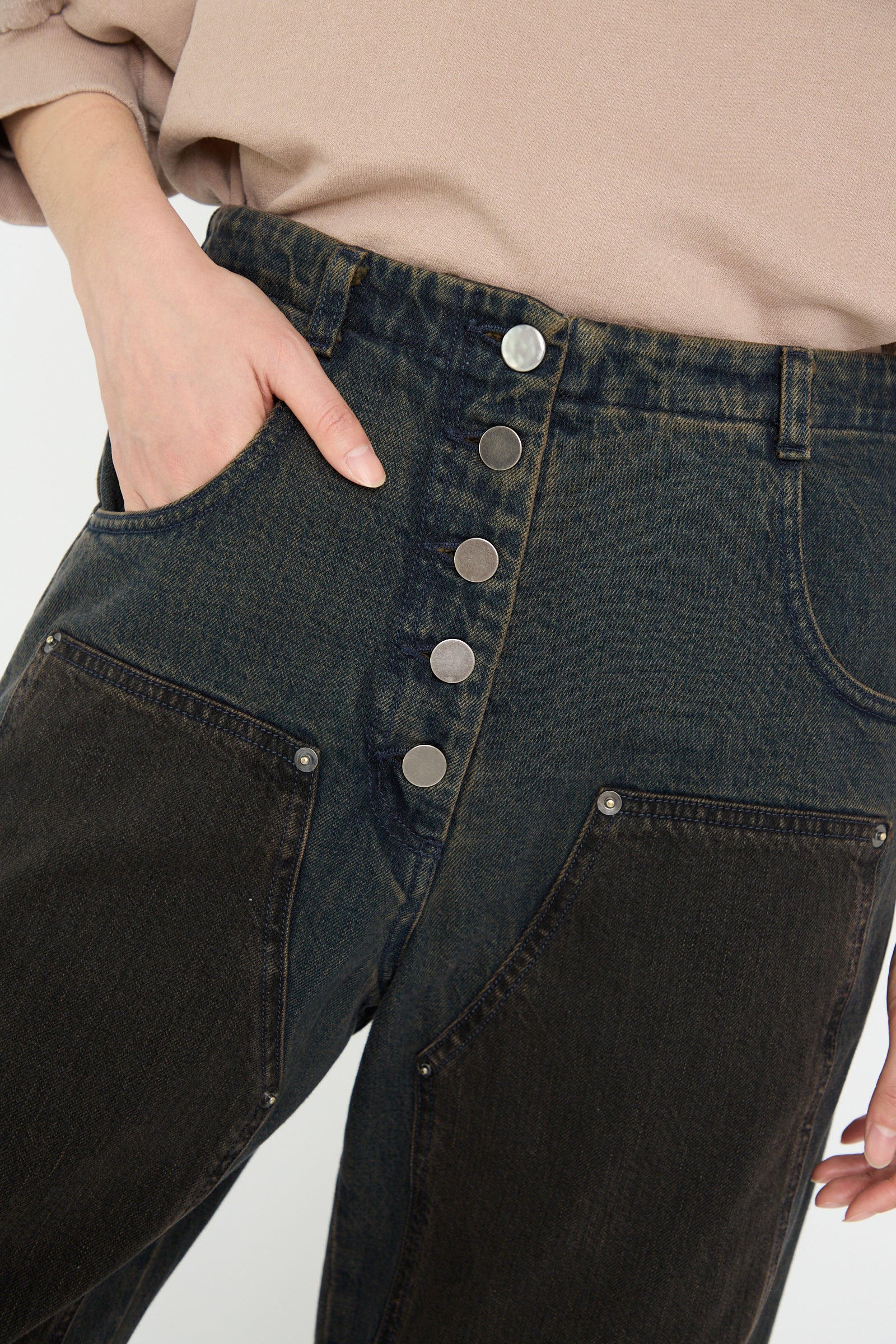 The image shows a close-up of someone wearing the Rachel Comey Handy Pant in brown, featuring relaxed-fit, made from dark Japanese denim. The pants have two front pockets and five large metallic buttons, with their left hand tucked into one pocket.