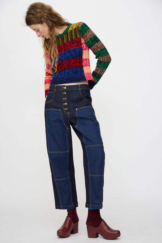 A person stands against a plain background wearing a multicolored striped sweater, Rachel Comey Handy Pant in Dark Indigo with a relaxed fit, and red clogs paired with maroon socks.