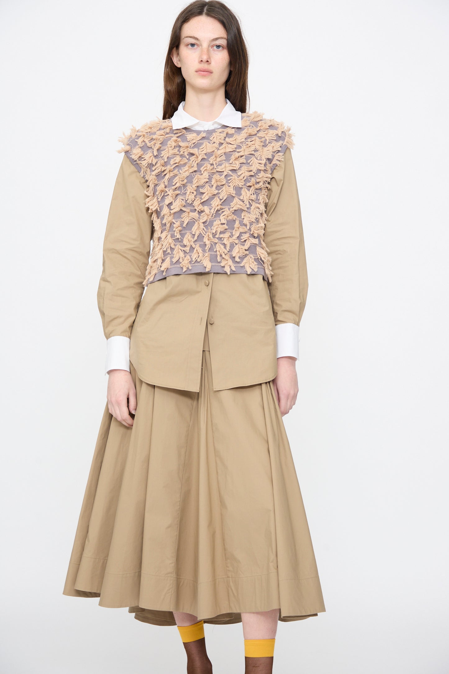A woman stands against a white background wearing the Rachel Comey Hazlitt Top in Dusty Mauve-Blush, styled with a beige button-up shirt and matching flared skirt.