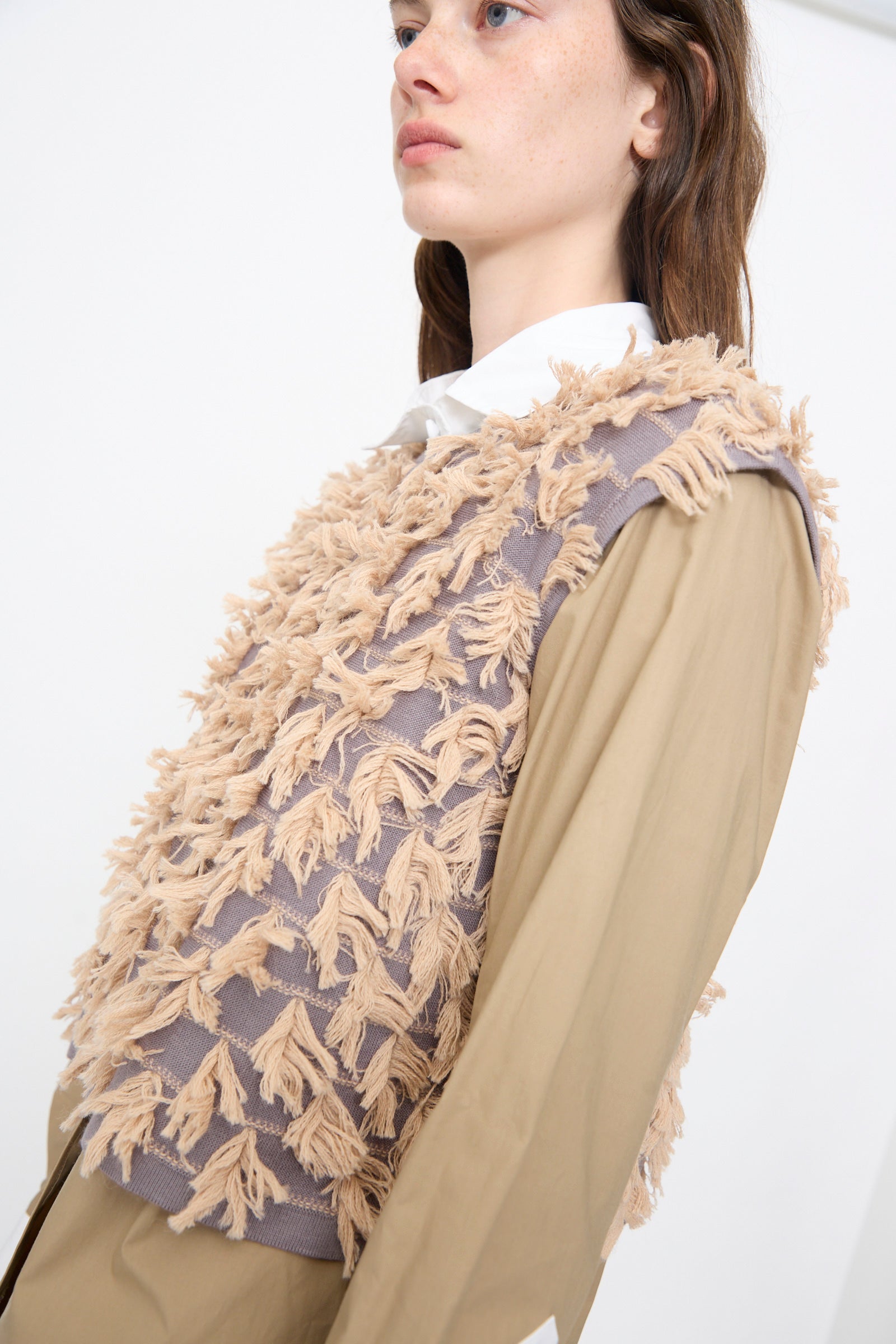 A person wears the Rachel Comey Hazlitt Top in Dusty Mauve-Blush, a sleeveless fringe top, over a tan long-sleeve shirt with a white collar, standing against a plain background.
