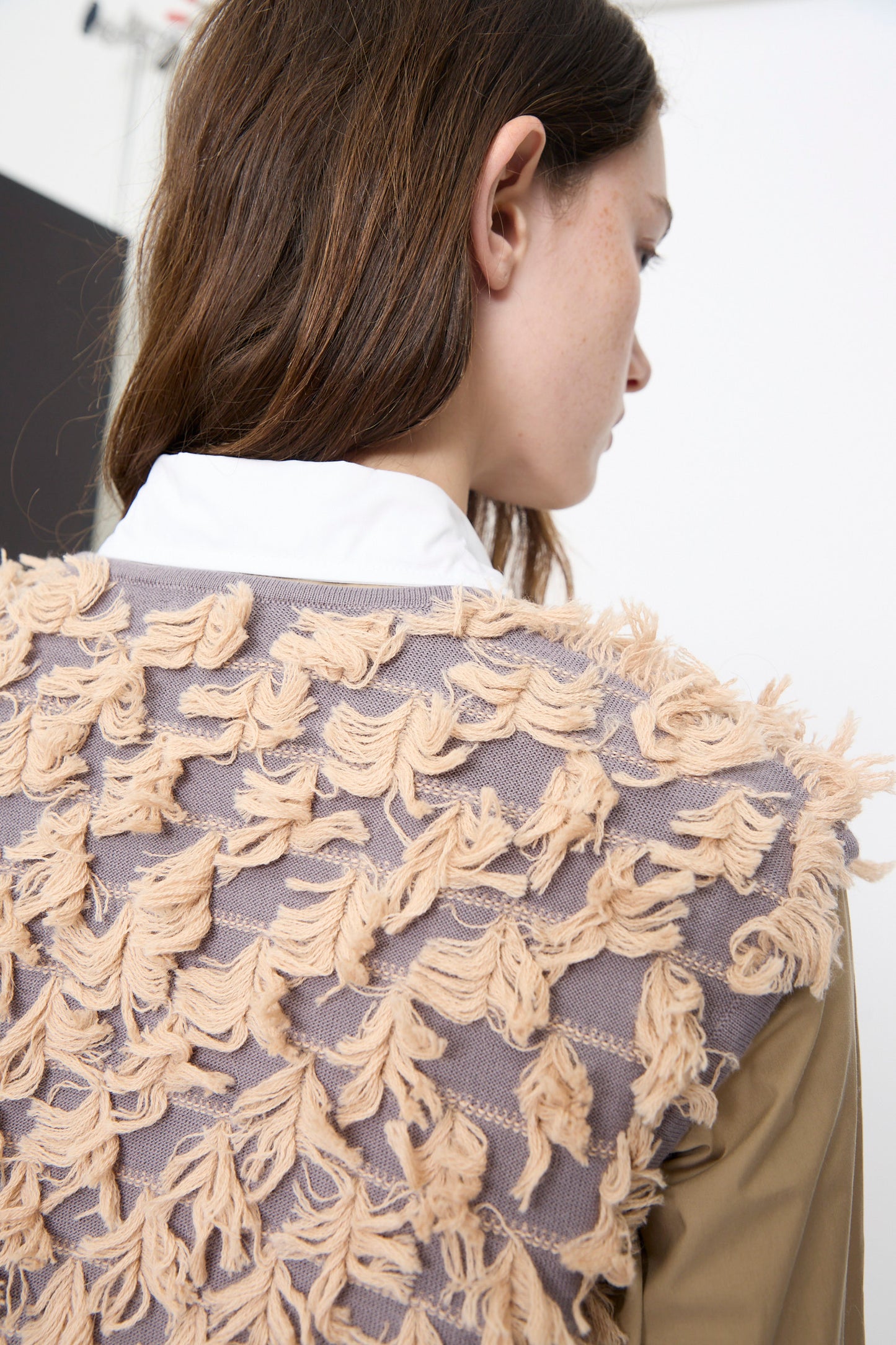 A person with long brown hair is seen from behind and to the side wearing the Rachel Comey Hazlitt Top in Dusty Mauve-Blush over a white collared shirt, paired with a tan jacket featuring textured beige fringe detailing.