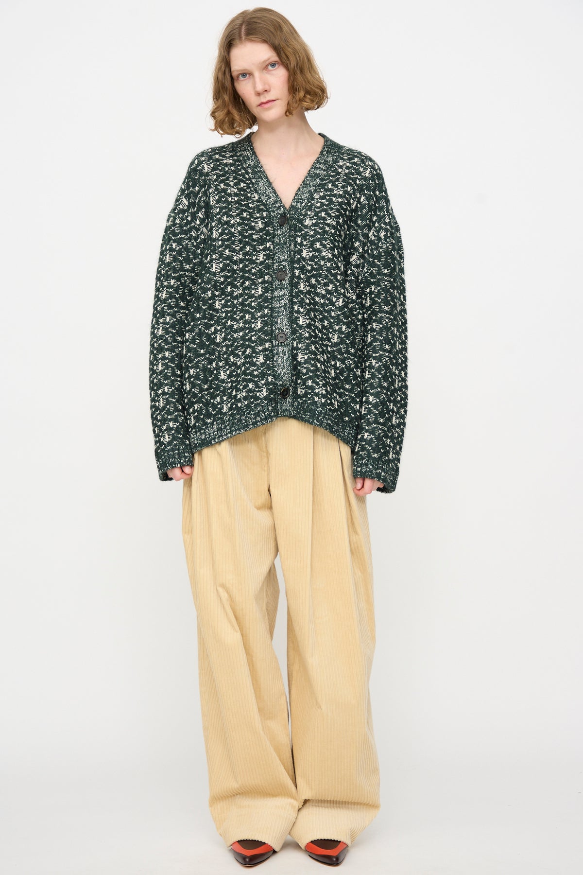 Wearing the Rachel Comey Hedda Cardigan in Forest—a loose dark green patterned cotton alpaca piece made in Peru—the person pairs it with wide beige corduroy pants and black shoes while standing against a plain white background.