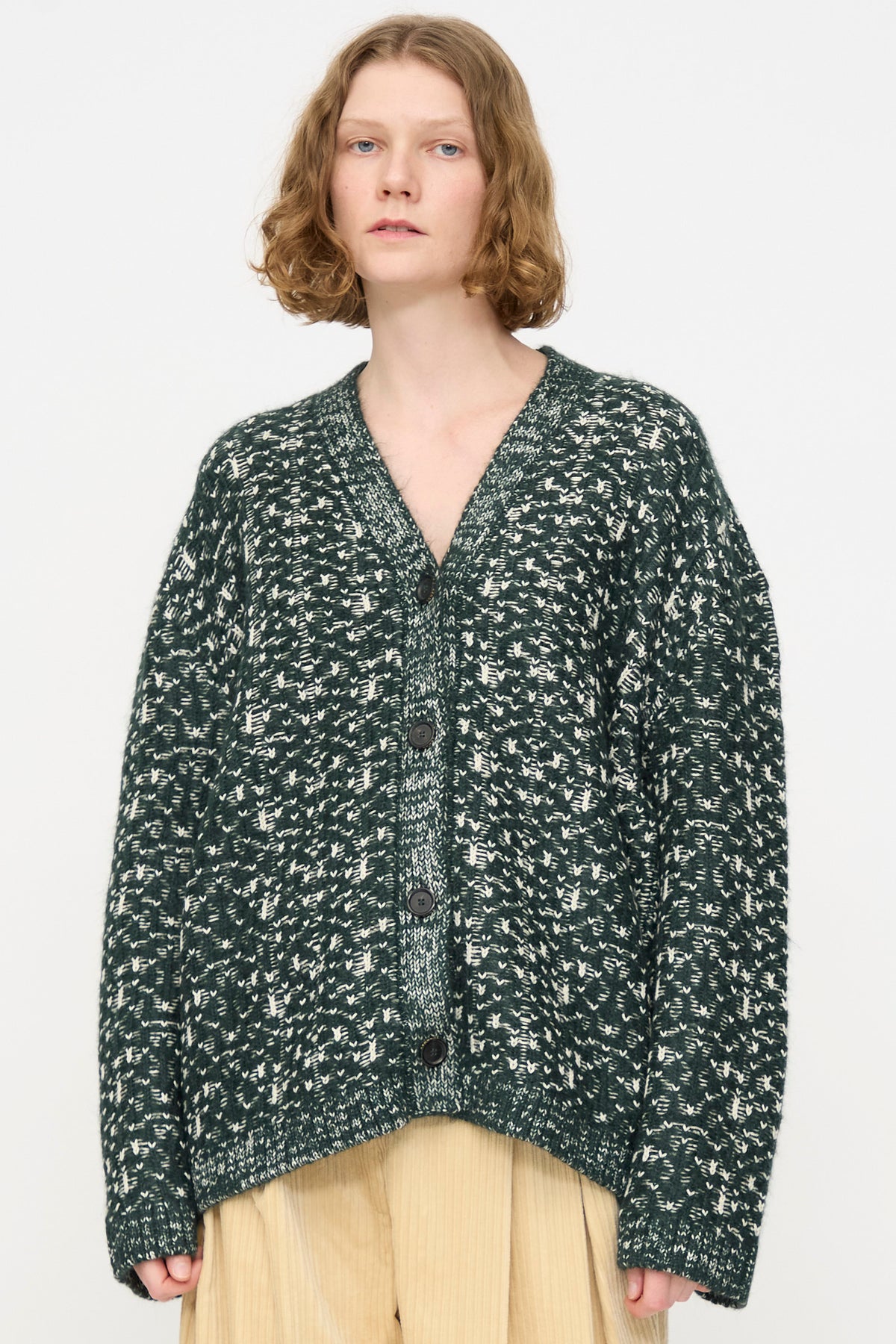 A person with short, wavy hair is wearing the Rachel Comey Hedda Cardigan in Forest—a loose, dark green and white patterned cotton alpaca cardigan with large buttons—paired with light-colored pants, standing against a plain white background.