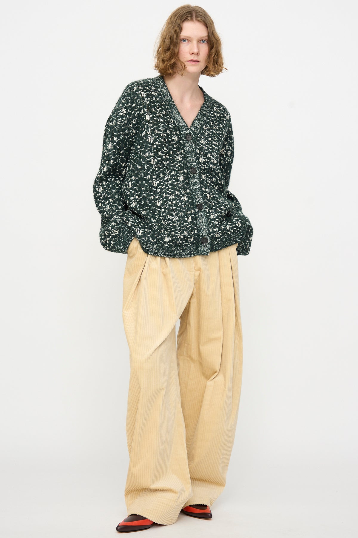 A person stands against a white background, wearing the Rachel Comey Hedda Cardigan in Forest—a dark green patterned merino wool piece made in Peru—paired with loose beige pleated pants and black and red shoes.