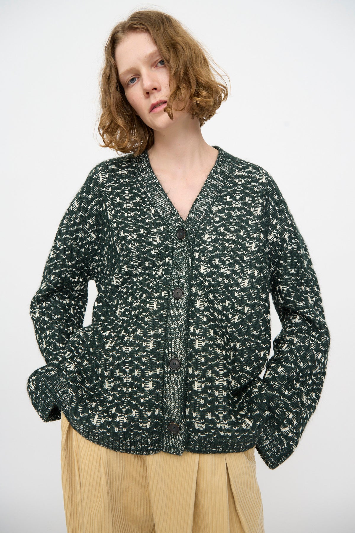 A person wearing the Rachel Comey Hedda Cardigan in Forest—a dark green patterned cotton alpaca button-up—paired with beige pleated pants, stands against a plain light background.