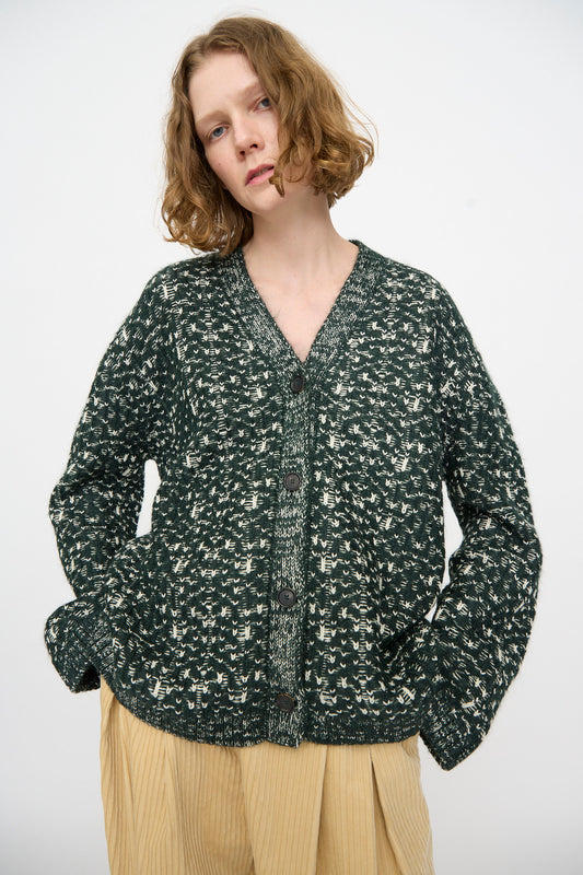 A person wearing the Rachel Comey Hedda Cardigan in Forest—a dark green patterned cotton alpaca button-up—paired with beige pleated pants, stands against a plain light background.