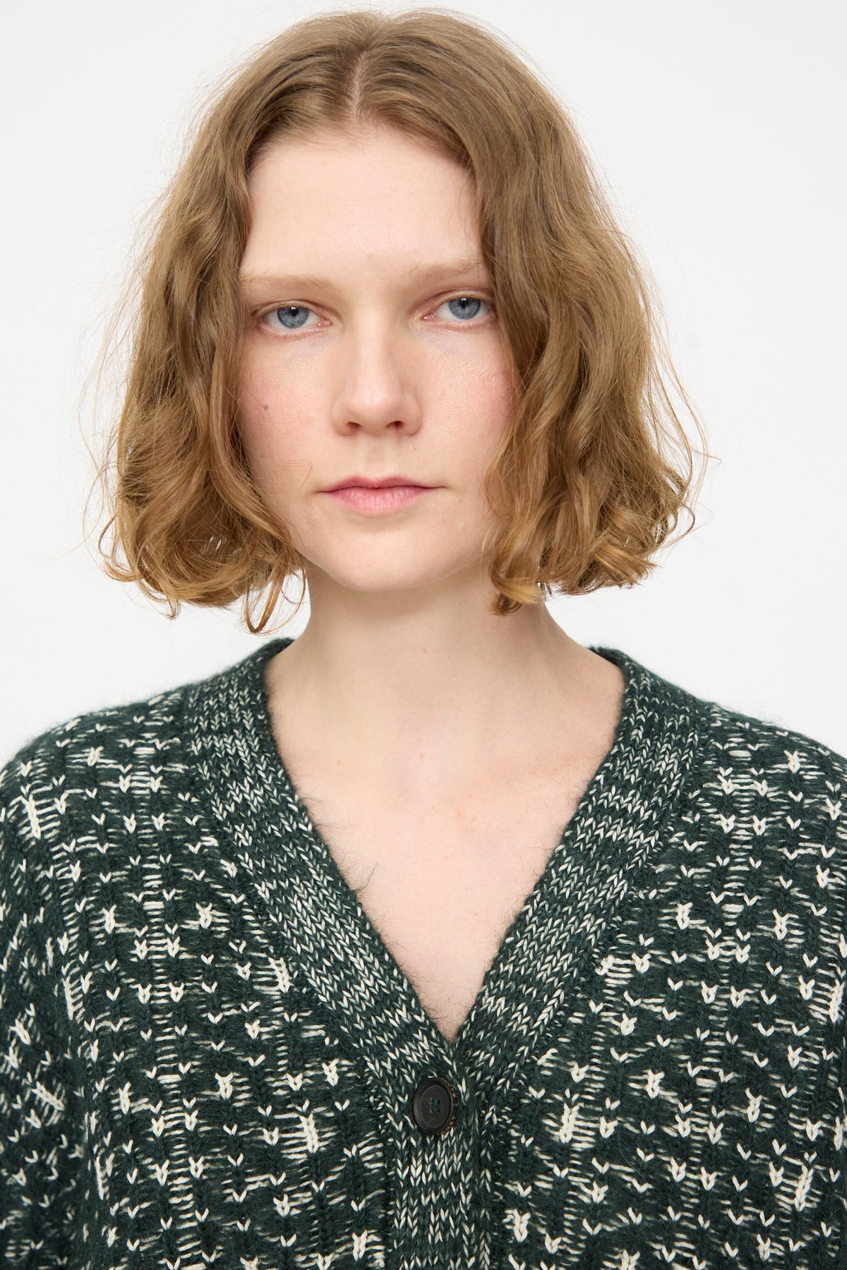 A person with short, wavy light brown hair is wearing the Rachel Comey Hedda Cardigan in Forest—a dark green and white patterned knit cotton-alpaca V-neck cardigan with button details, made in Peru—standing against a plain white background.