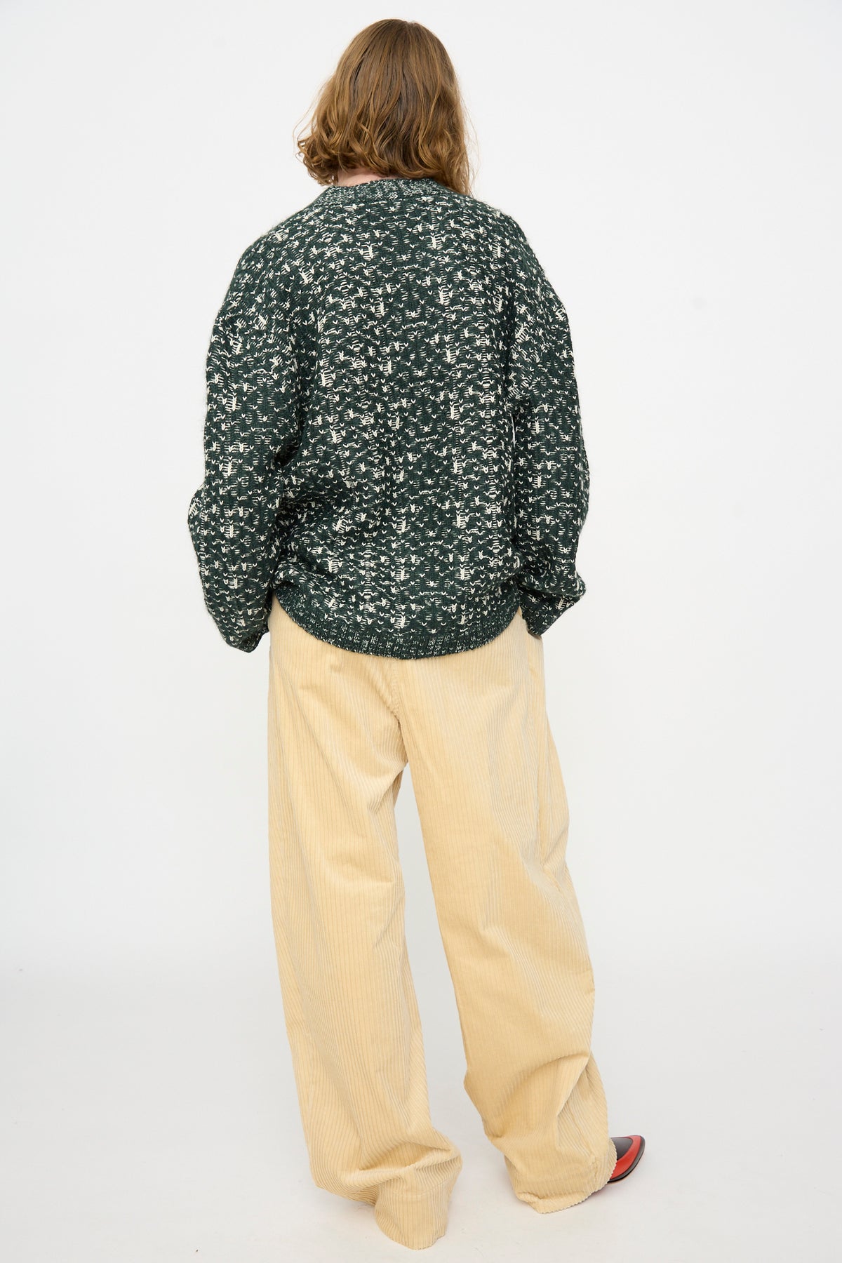 A person stands with their back to the camera, wearing the Rachel Comey Hedda Cardigan in Forest—a dark green patterned merino wool piece made in Peru—paired with loose beige corduroy pants and dark shoes, set against a plain white background.