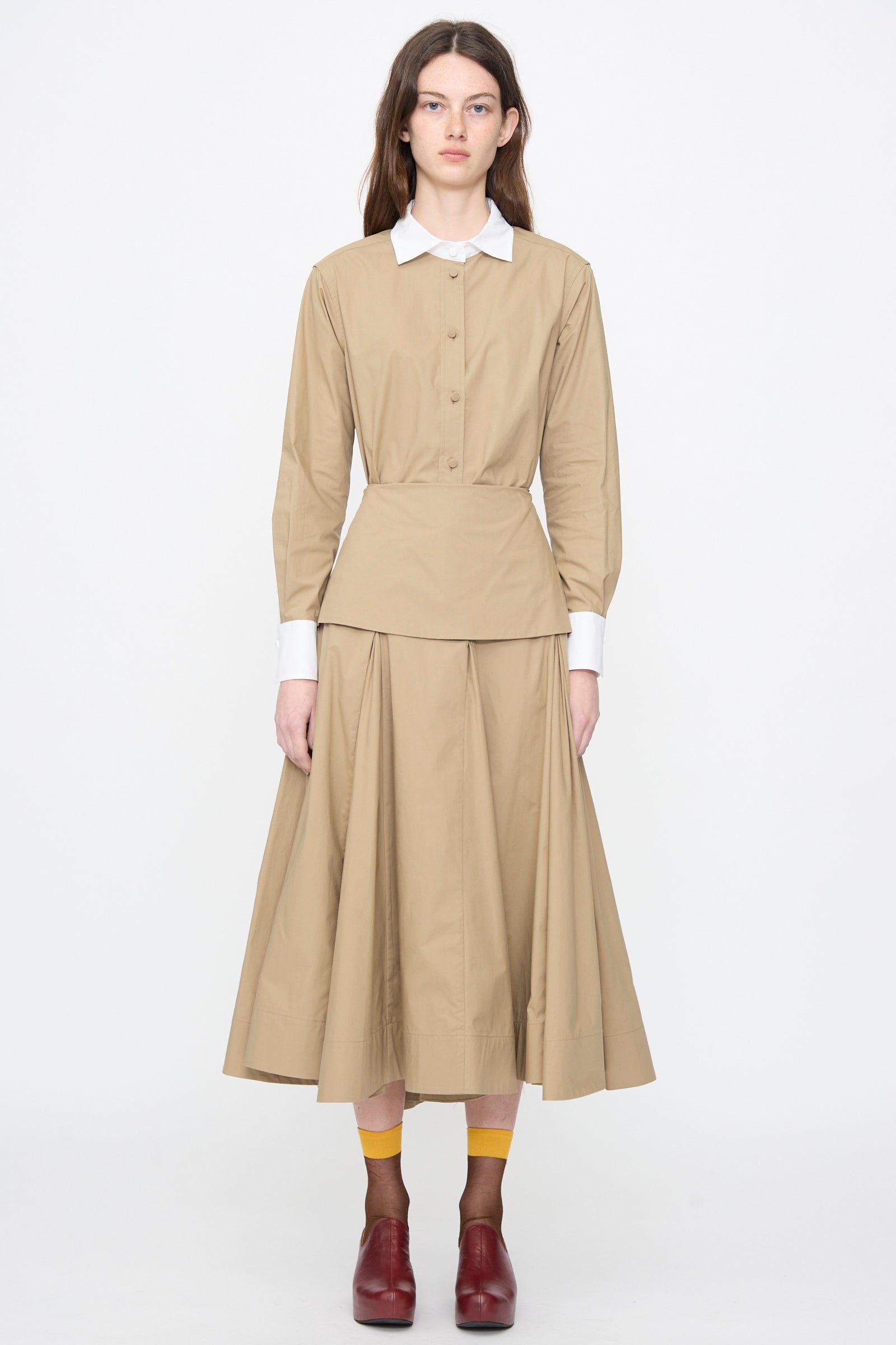A person stands facing forward wearing the Rachel Comey Heni Skirt in Khaki, styled with a coordinating beige blouse, yellow socks, and brown shoes, set against a plain white background.