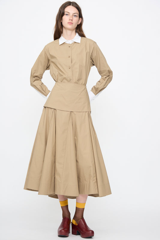 A woman stands against a plain background wearing the Rachel Comey Heni Skirt in Khaki, paired with a tan long-sleeve shirt with a white collar, yellow socks, and dark red shoes.