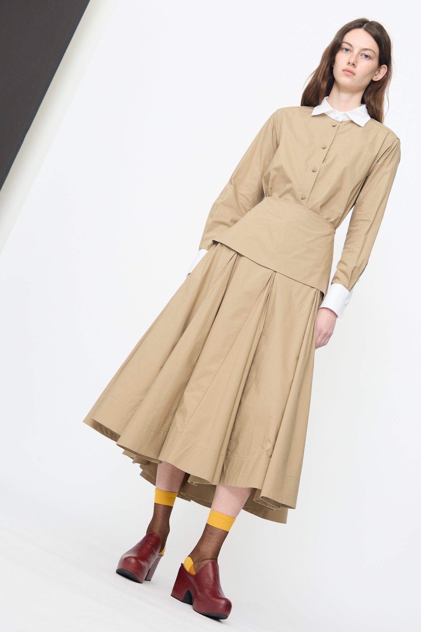 A woman wearing the Rachel Comey Heni Skirt in Khaki with a beige long-sleeve blouse, white collar and cuffs, yellow socks, and red platform shoes stands against a plain white background.