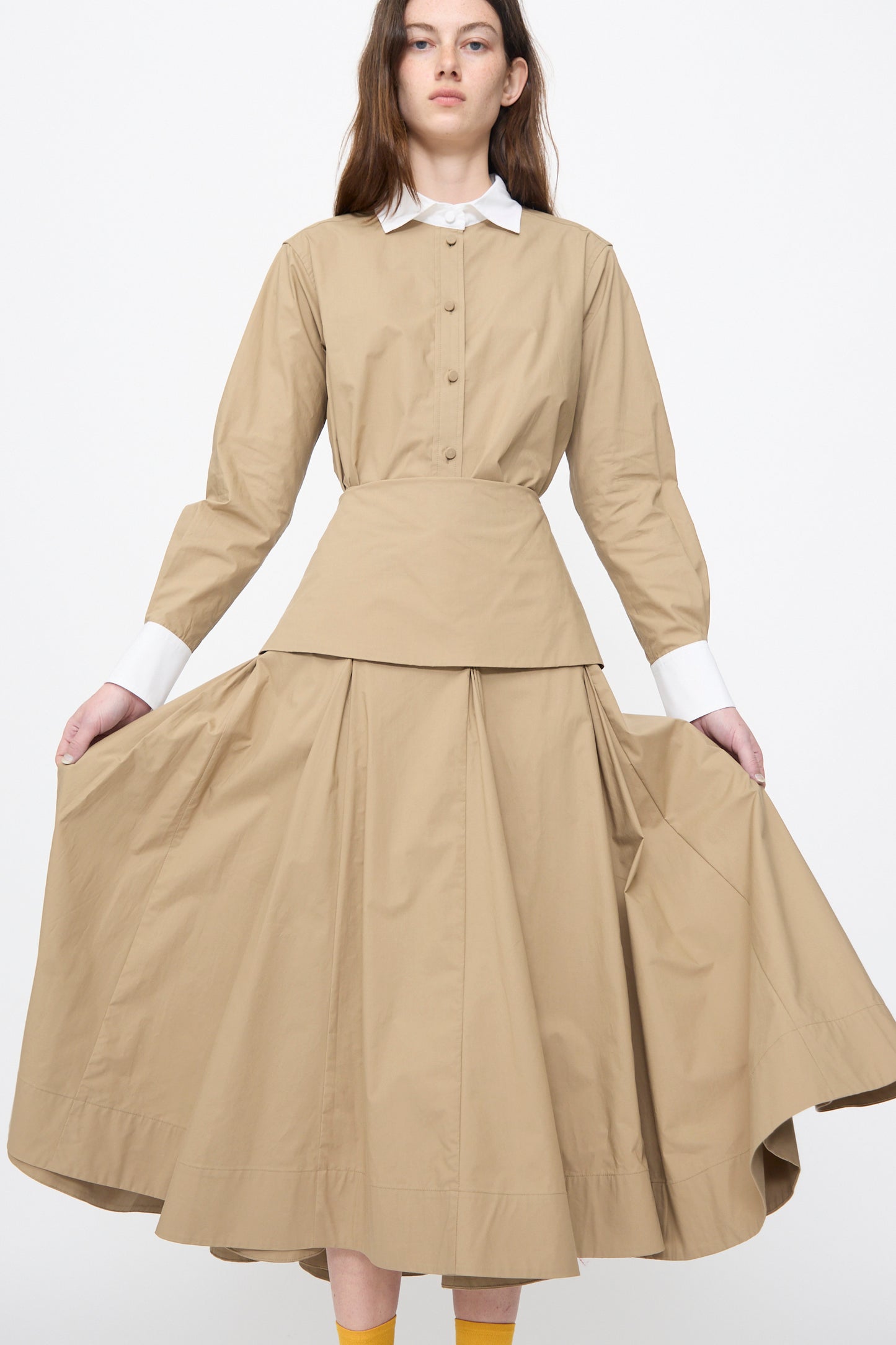 A woman stands against a white background wearing the Rachel Comey Heni Skirt in Khaki, paired with a beige cotton poplin long-sleeve button-up top featuring white cuffs and collar.