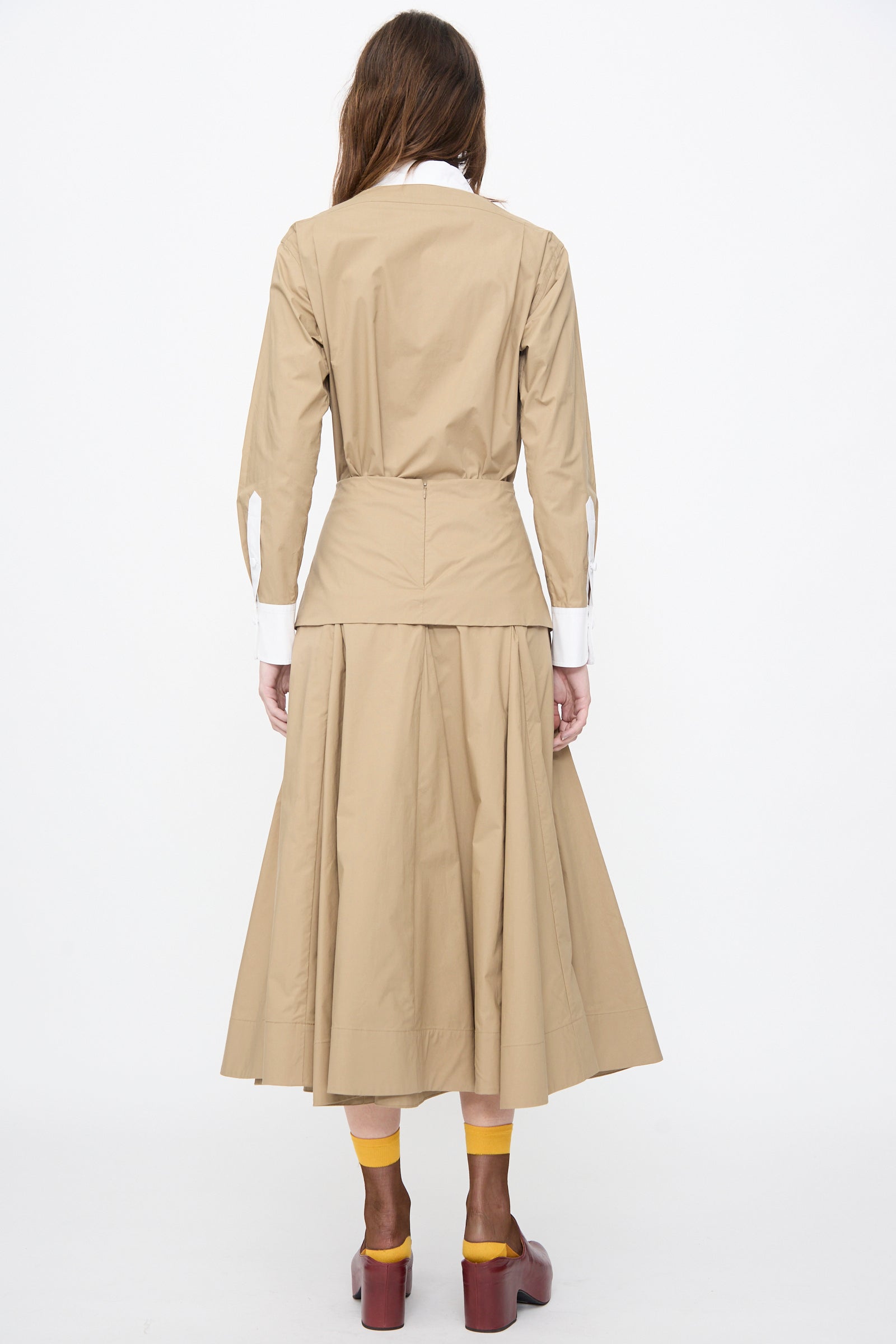 A woman stands with her back to the camera, wearing the Rachel Comey Heni Skirt in Khaki paired with a tan cotton poplin long-sleeve top, yellow socks, and brown platform shoes against a plain white background.