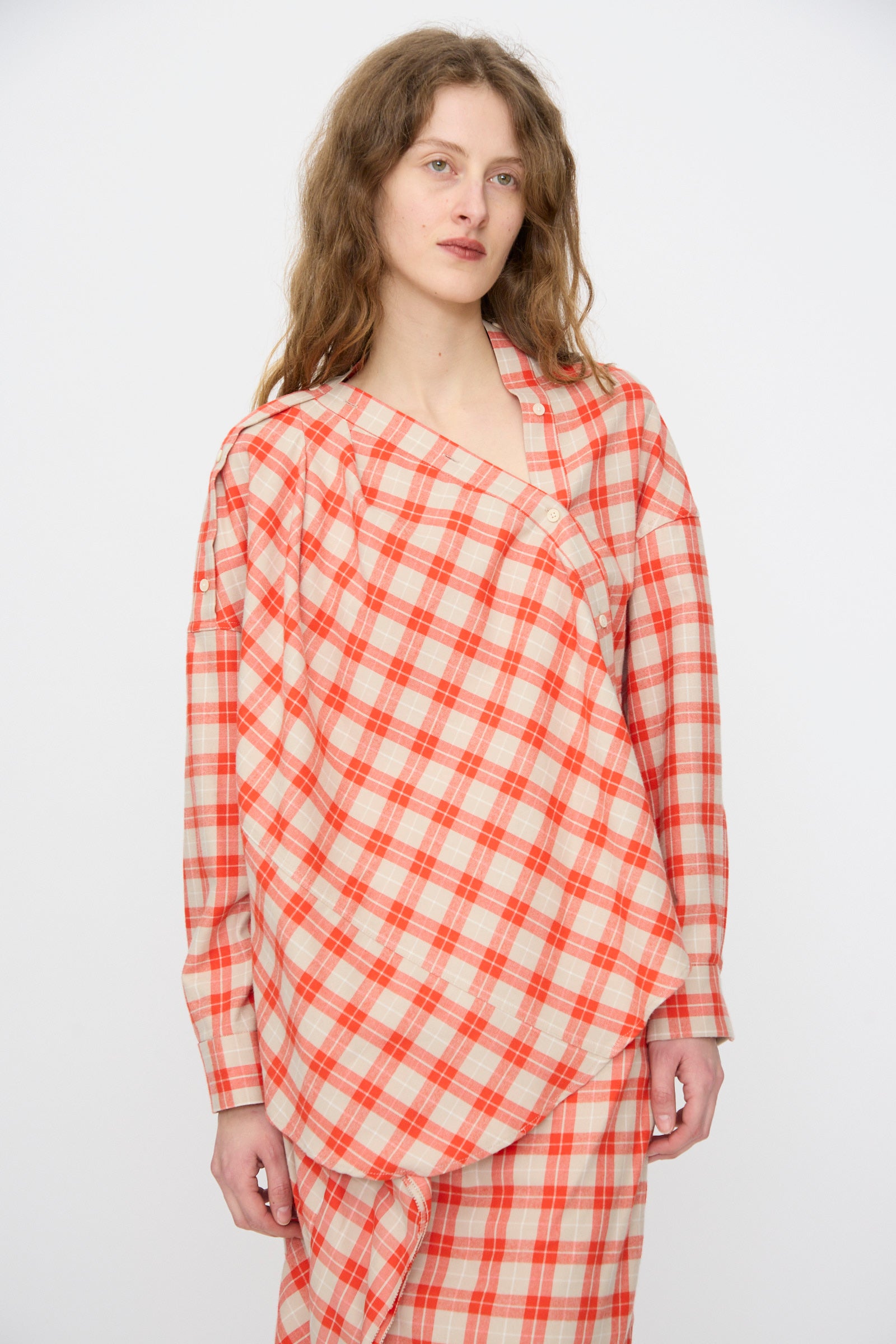 A person with long wavy hair wears the Rachel Comey Hollow Top in Red, a red and beige plaid cotton flannel piece with an asymmetrical draped design, paired with a matching skirt, standing against a plain light background.