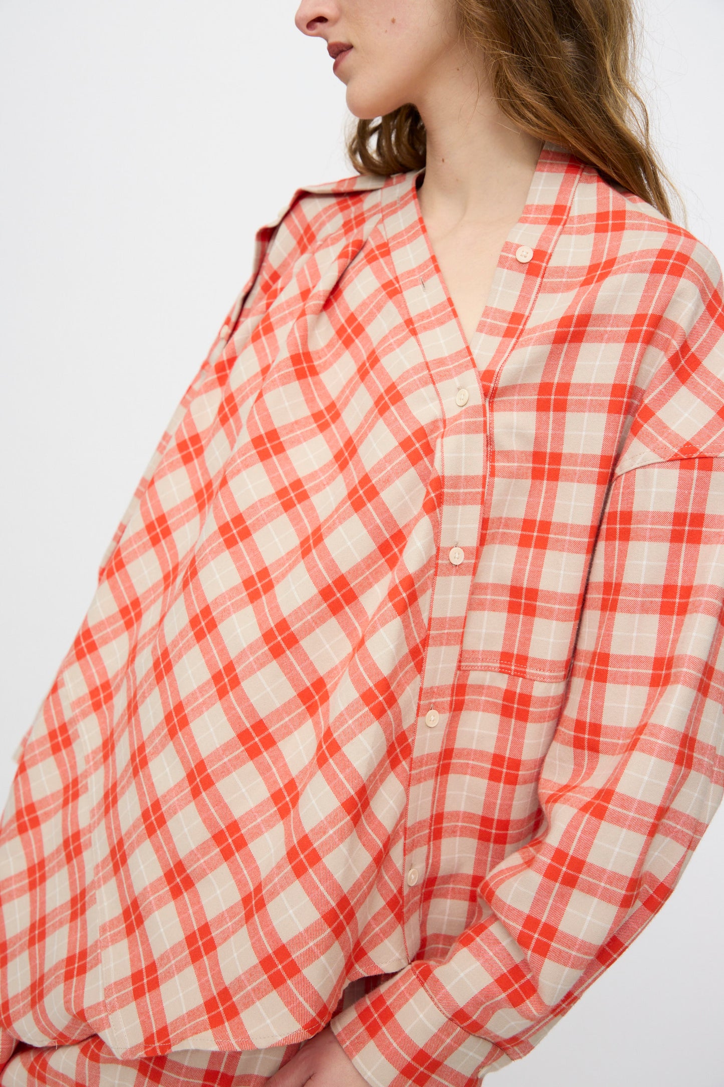 A woman stands against a plain white background, wearing the Rachel Comey Hollow Top in Red—a loose-fitting cotton flannel with red and beige plaid and button detail.