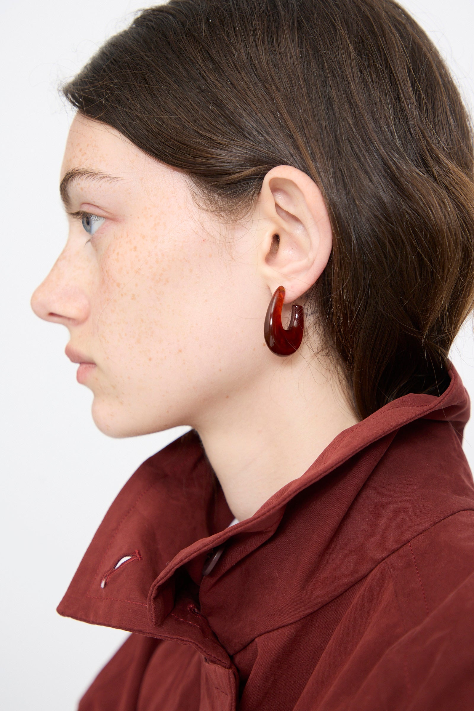 A person with fair skin and brown hair wears a maroon coat and the Rachel Comey Isle Earring in Dark Tortoise, shown in side profile against a plain background.