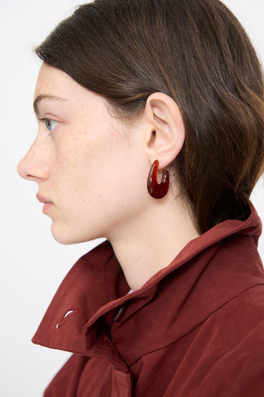 A person with fair skin and brown hair wears a maroon coat and the Rachel Comey Isle Earring in Dark Tortoise, shown in side profile against a plain background.