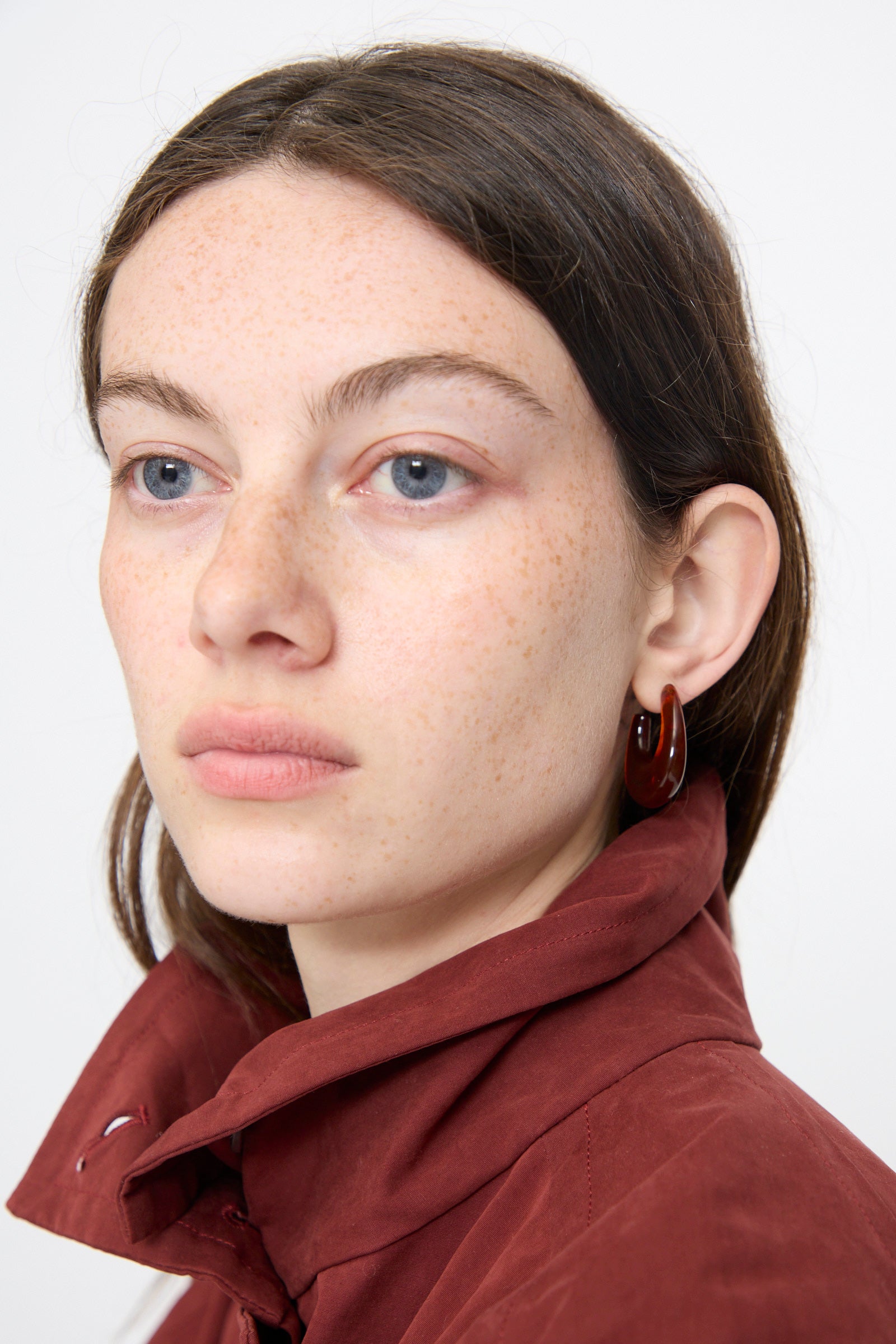 A light-skinned person with blue eyes and brown hair wears a maroon jacket and the Rachel Comey Isle Earring in Dark Tortoise, gazing slightly to the side against a plain background.