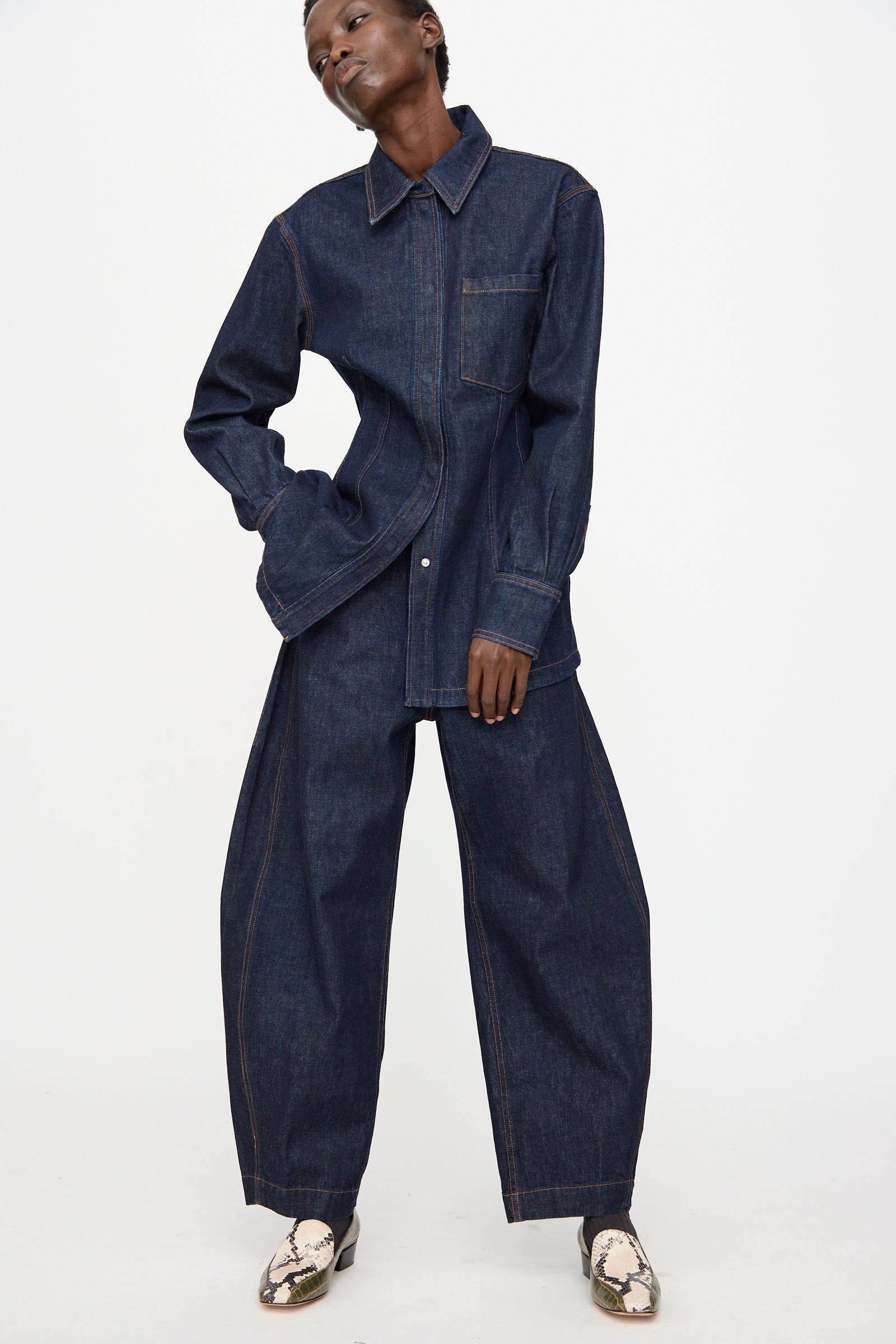 A person is wearing the Rachel Comey Keenan Pant in Raw Wash, paired with a loose dark denim shirt and light snakeskin shoes, standing against a plain white background.