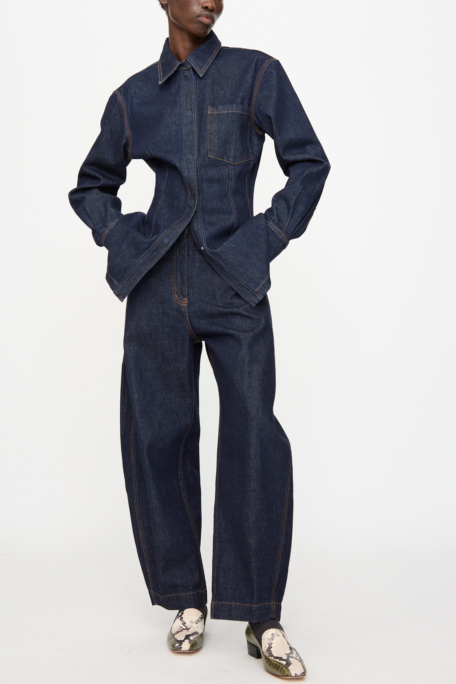 A person wears the Rachel Comey Keenan Pant in Raw Wash, featuring high waist barrel legs and visible stitching, styled with light snakeskin-patterned shoes, against a plain white background.