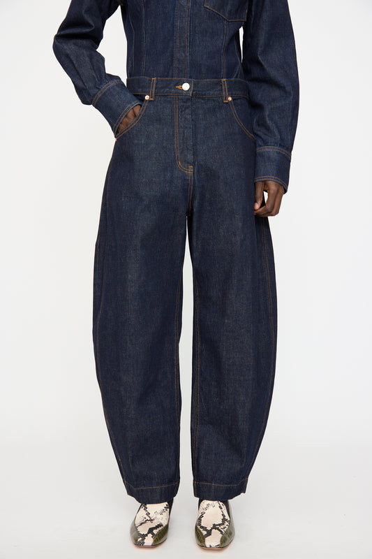 A person wears the Rachel Comey Keenan Pant in Raw Wash, a high waist barrel leg dark denim, paired with tan snake print boots, standing against a plain white background.