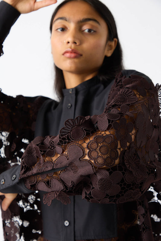 A person wearing the Rachel Comey Lace Flower Myra Top in Dark Brown and Black, featuring intricate crochet lace patterns on the sleeves, stands against a plain background. One arm is raised, partially covering the face.