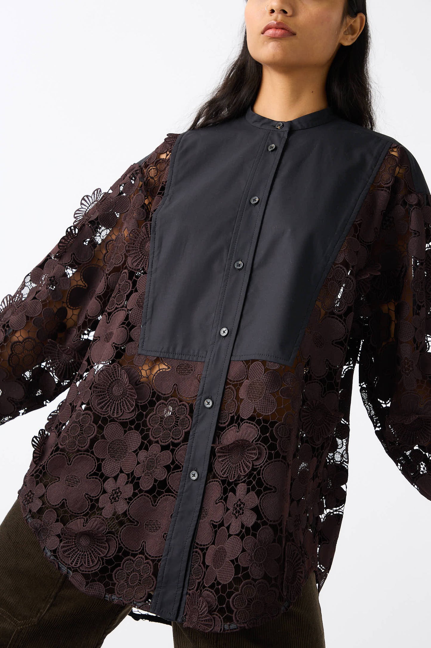 A person is wearing the Rachel Comey Lace Flower Myra Top in black, which features 3D floral appliques on the lace sleeves and a buttoned front panel. The image captures the upper body and part of the legs, showcasing its relaxed fit.