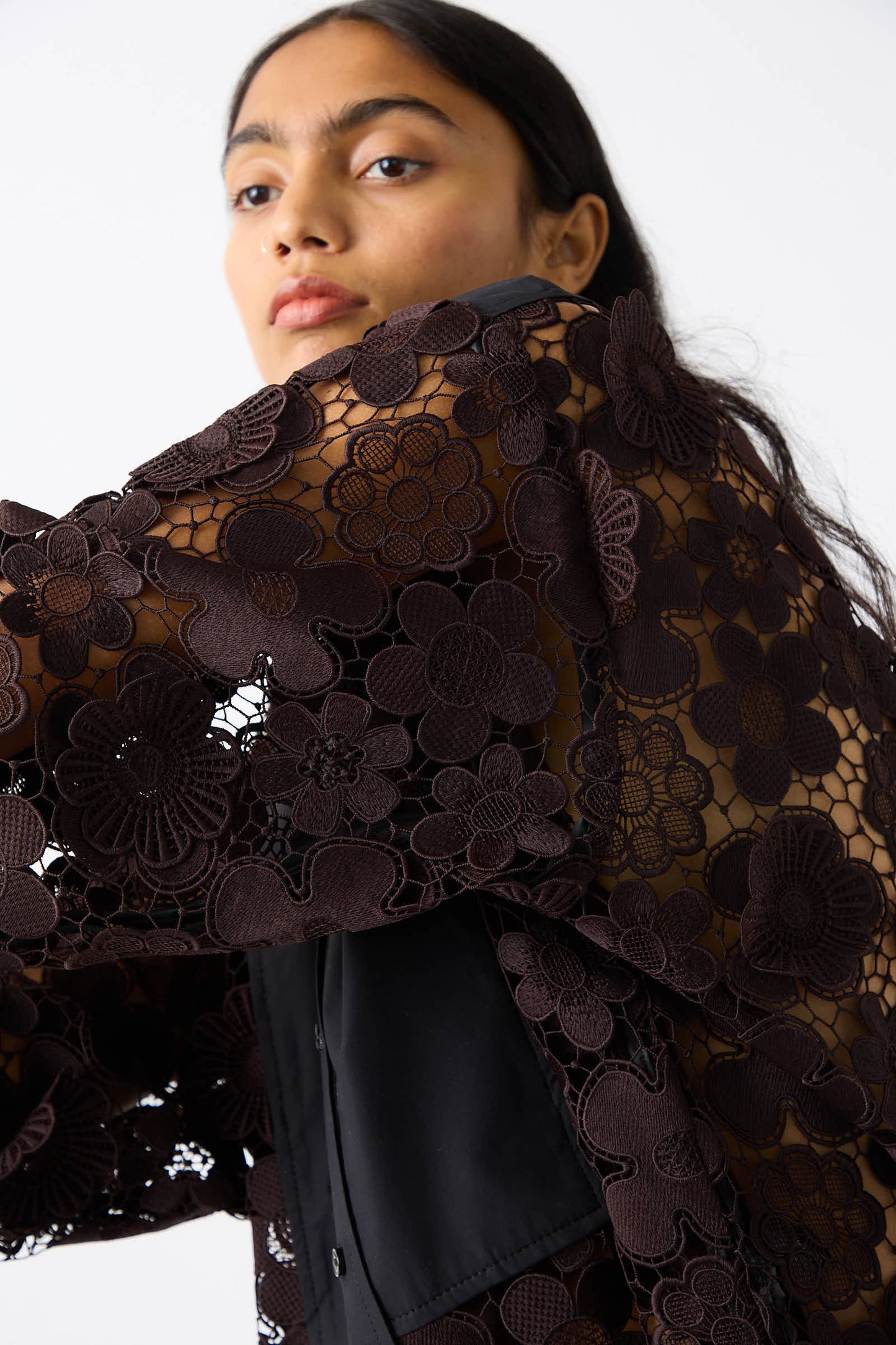 A person with long dark hair wears the Lace Flower Myra Top in Dark Brown and Black by Rachel Comey, featuring crochet lace details and 3D floral appliques, looking to the side against a white background.