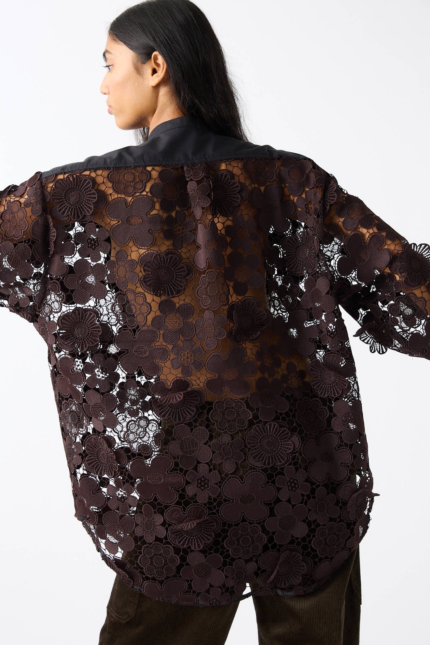 Person wearing a relaxed fit Rachel Comey Lace Flower Myra Top in black sheer lace, adorned with crochet lace and 3D floral appliques, facing away from the camera, with long dark hair and arms stretched out.