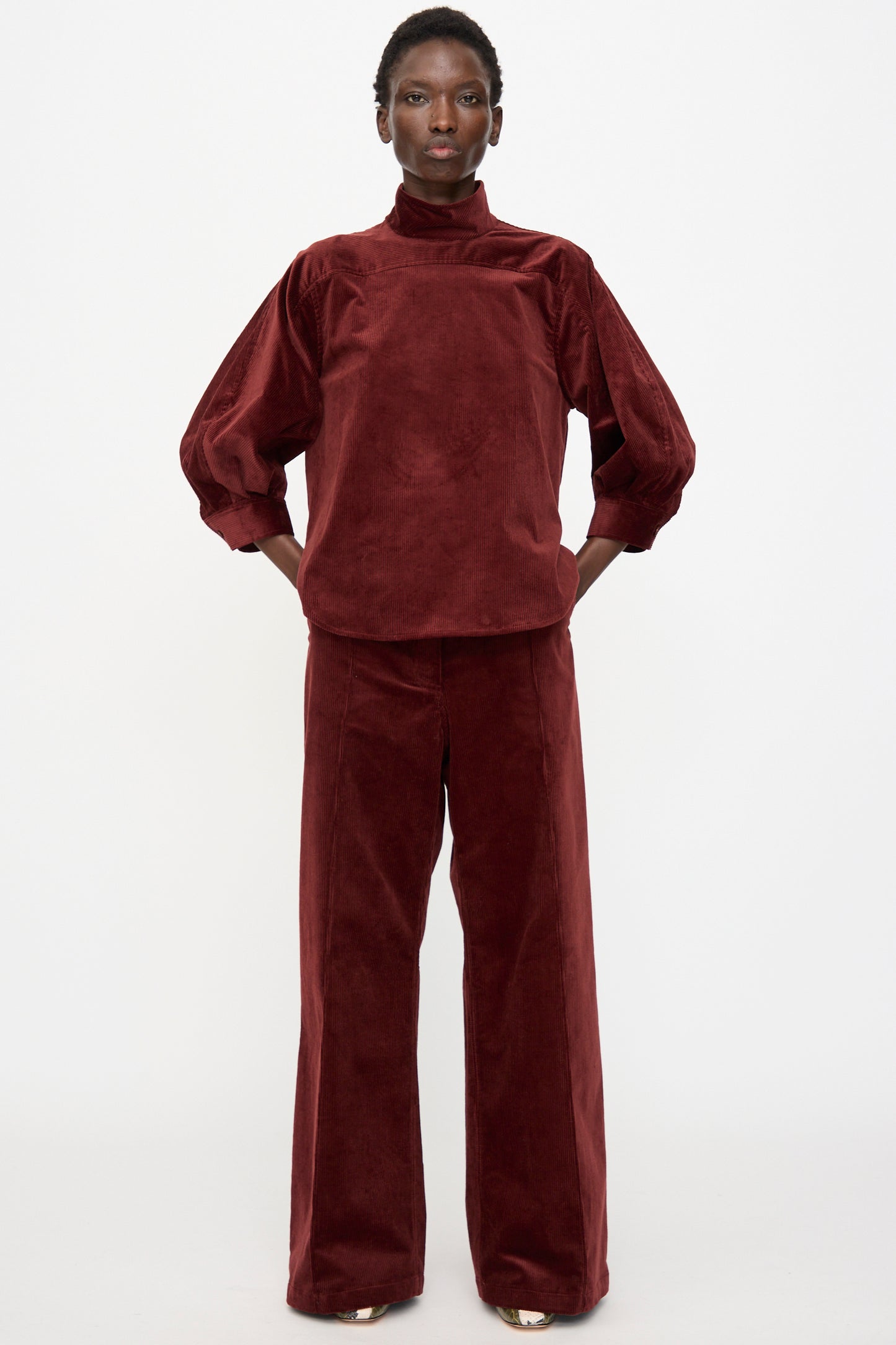 A person stands against a plain white background wearing a burgundy velvet high-neck, long-sleeve top paired with Rachel Comey's Lanett Cord Pant in Brick.