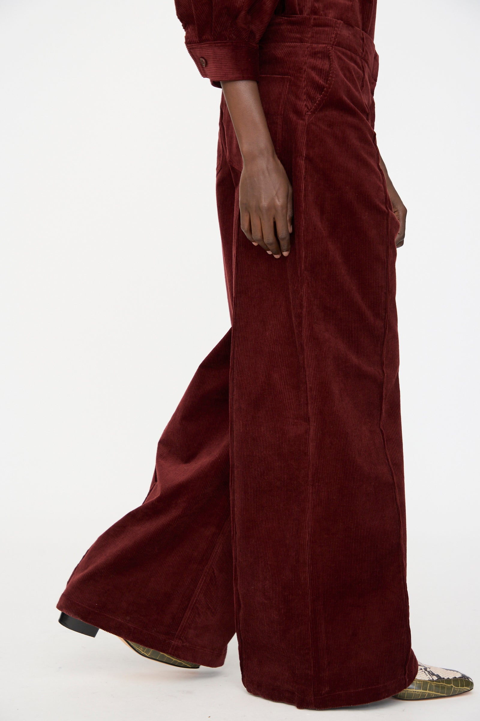 A person stands against a white background wearing Rachel Comey’s Lanett Cord Pant in Brick with brown pointed-toe shoes.