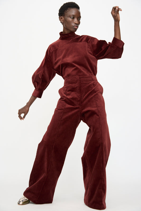 A model poses with one arm raised against a white background, wearing the Rachel Comey Lanett Cord Pant in Brick and a burgundy velvet top.