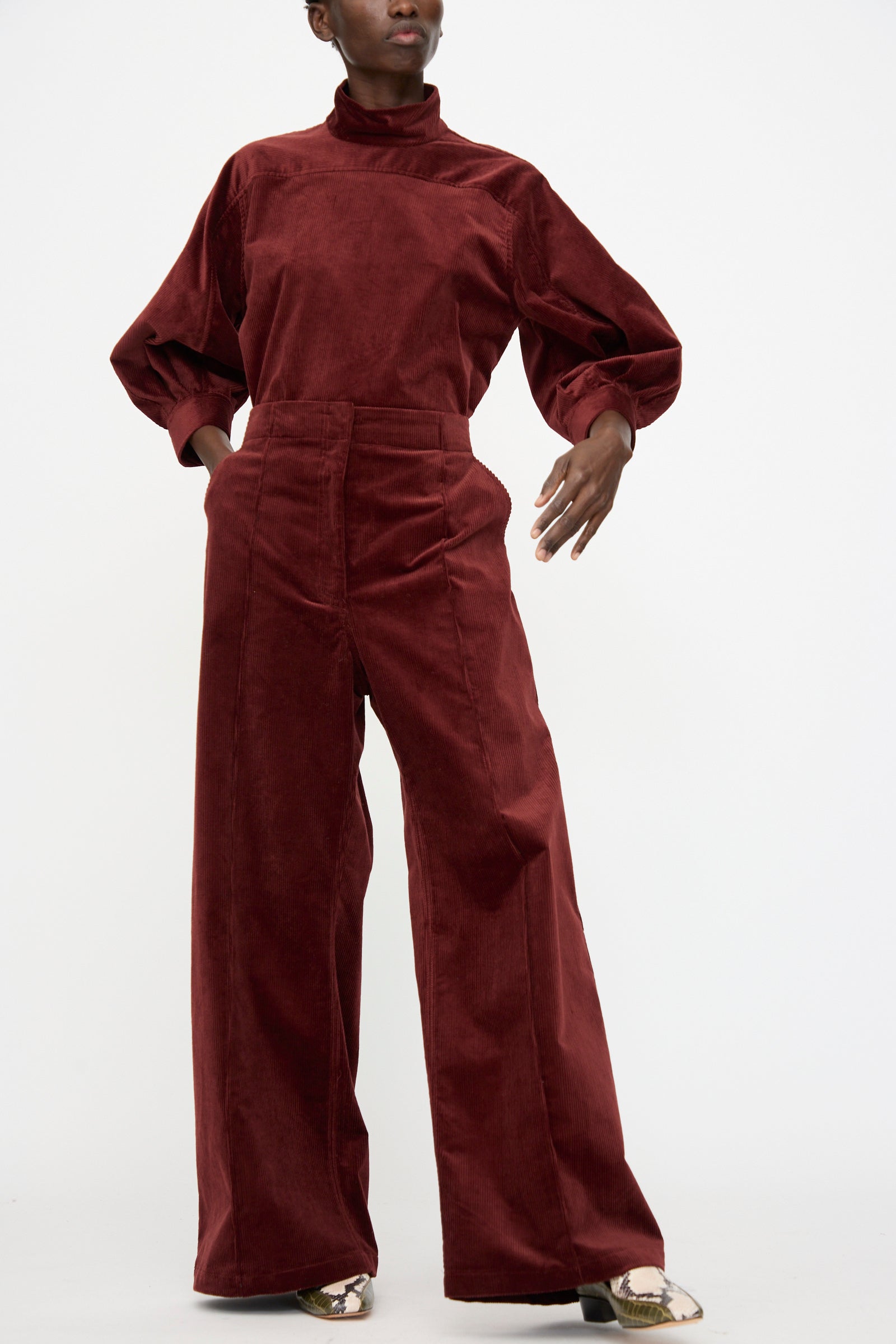 A person wears the Rachel Comey Lanett Cord Pant in Brick, paired with a matching burgundy turtleneck top, standing against a plain white background.