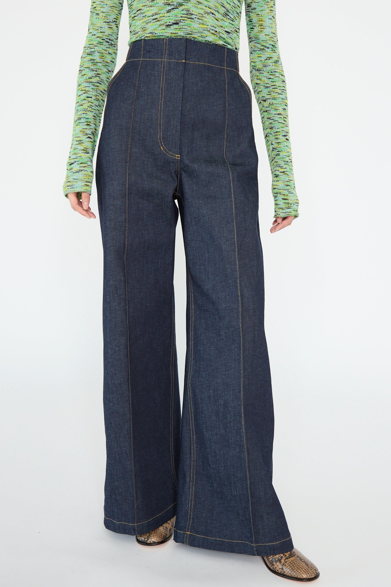 A model is wearing the Rachel Comey Lanett Pant in Super Raw, high-waisted wide-leg denim, with a green patterned long-sleeve top and brown shoes, against a white backdrop. 