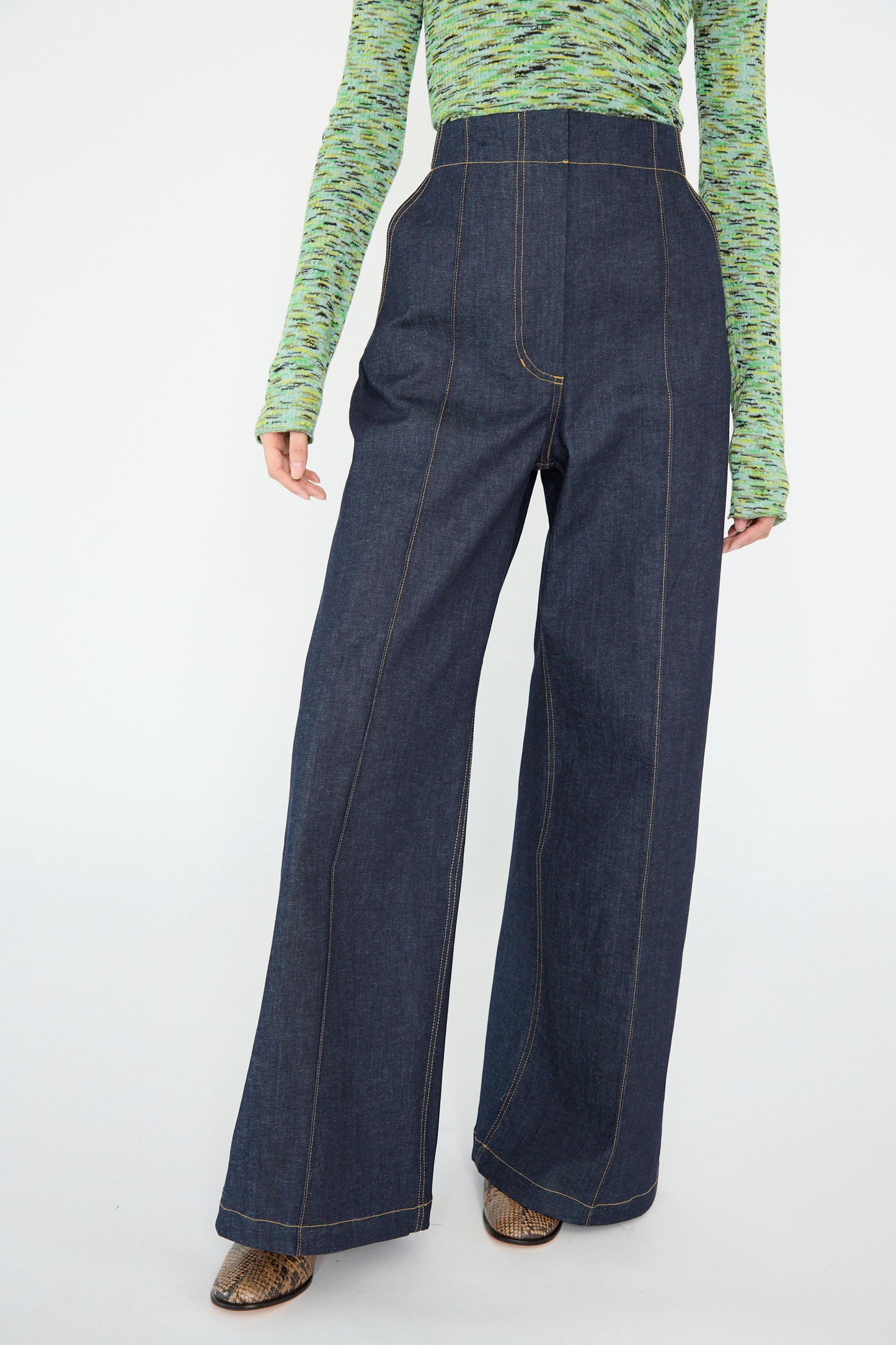 A model wearing Rachel Comey's Lanett Pant in Super Raw denim with a green patterned long-sleeve top, partially revealing brown ankle boots, against a white backdrop. 