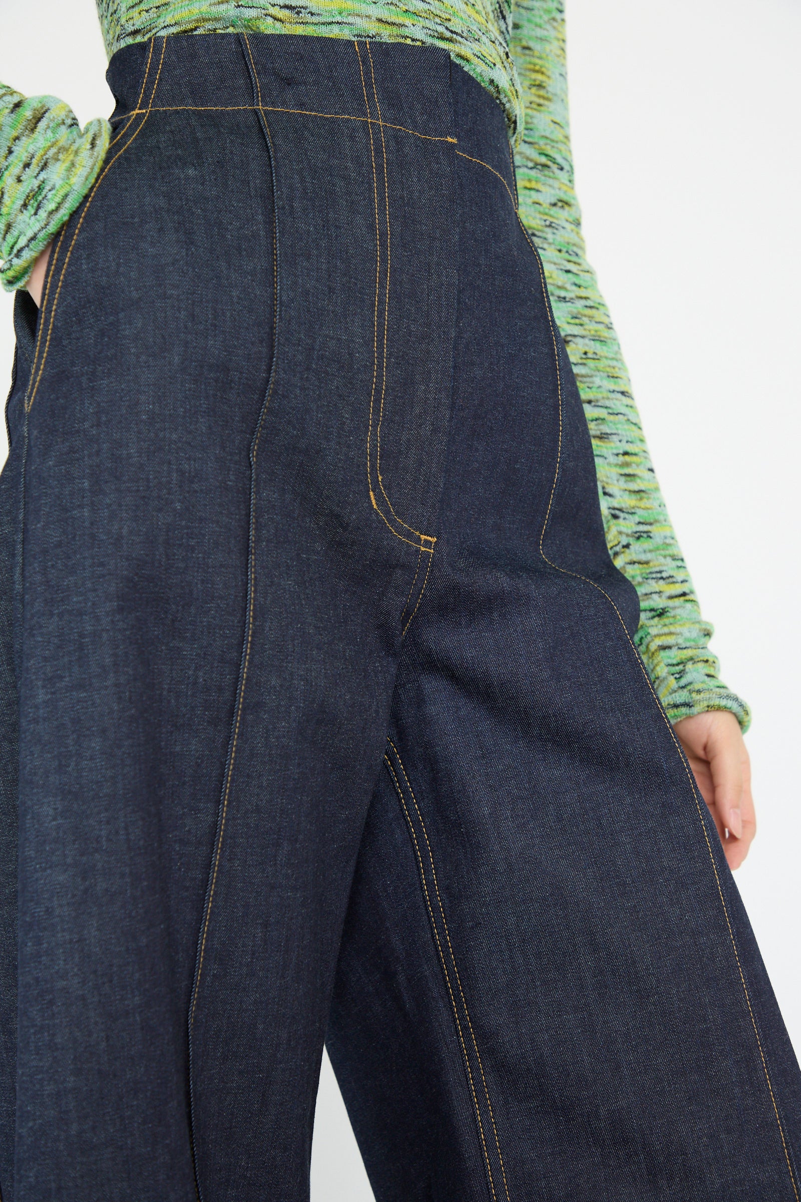 A close-up of a person wearing Rachel Comey's Lanett Pant in Super Raw, featuring high-waisted dark denim with visible stitching, paired with a green textured knit top.