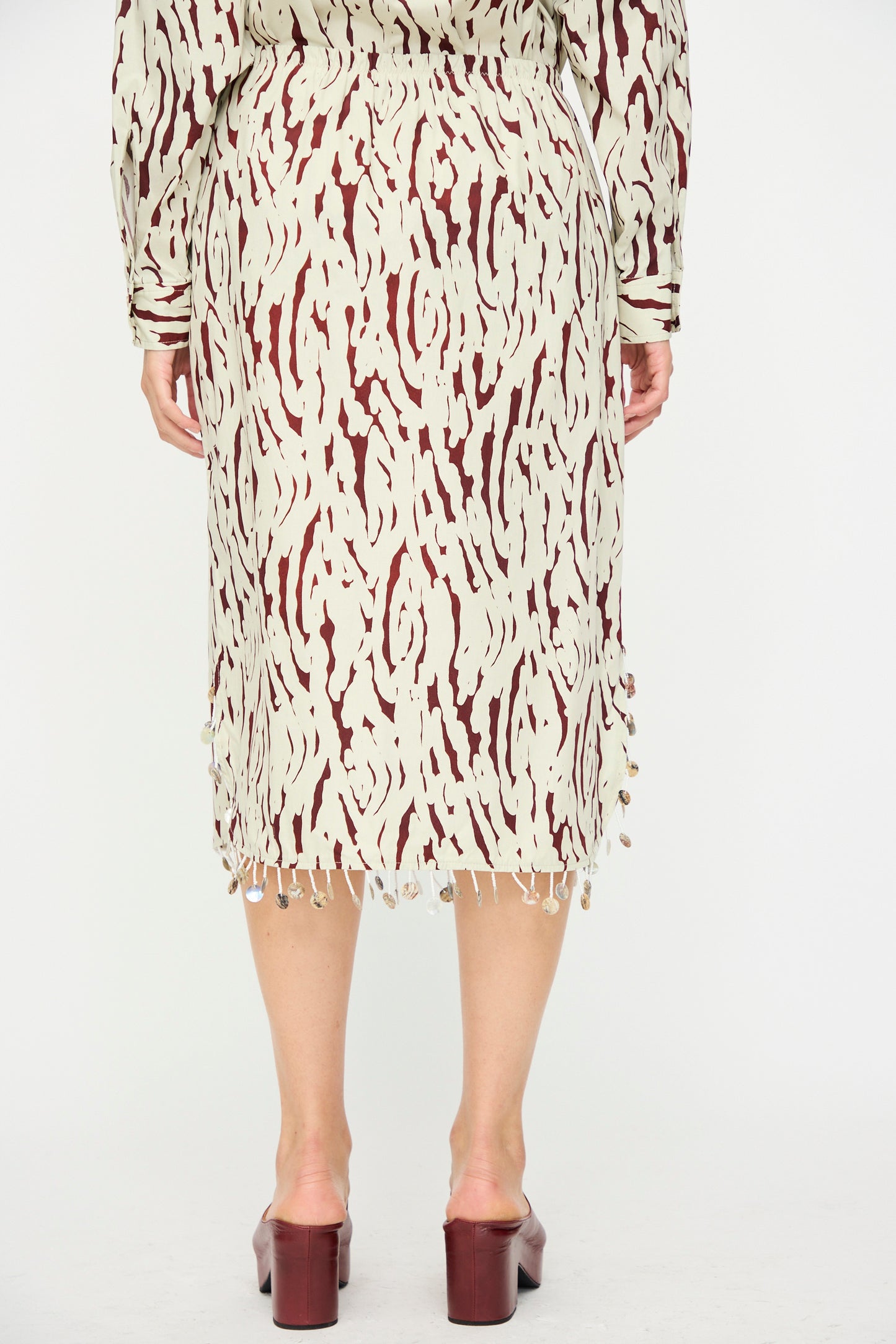 A person wears the Rachel Comey Lastica Skirt in Cream, featuring an abstract pattern and fringe, paired with maroon heeled sandals. Photographed from the back against a white background.