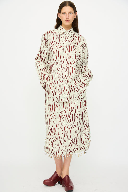 A person stands against a white background wearing the Rachel Comey Lastica Skirt in Cream with an abstract print, paired with a loose cream and burgundy shirt and burgundy platform shoes.