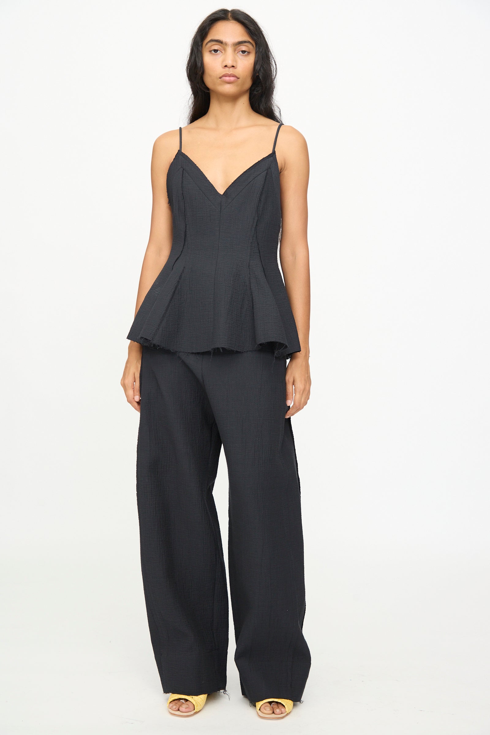 A woman stands against a plain background, wearing the Rachel Comey Layley Top in Black, paired with wide-leg black pants and open-toe yellow shoes.