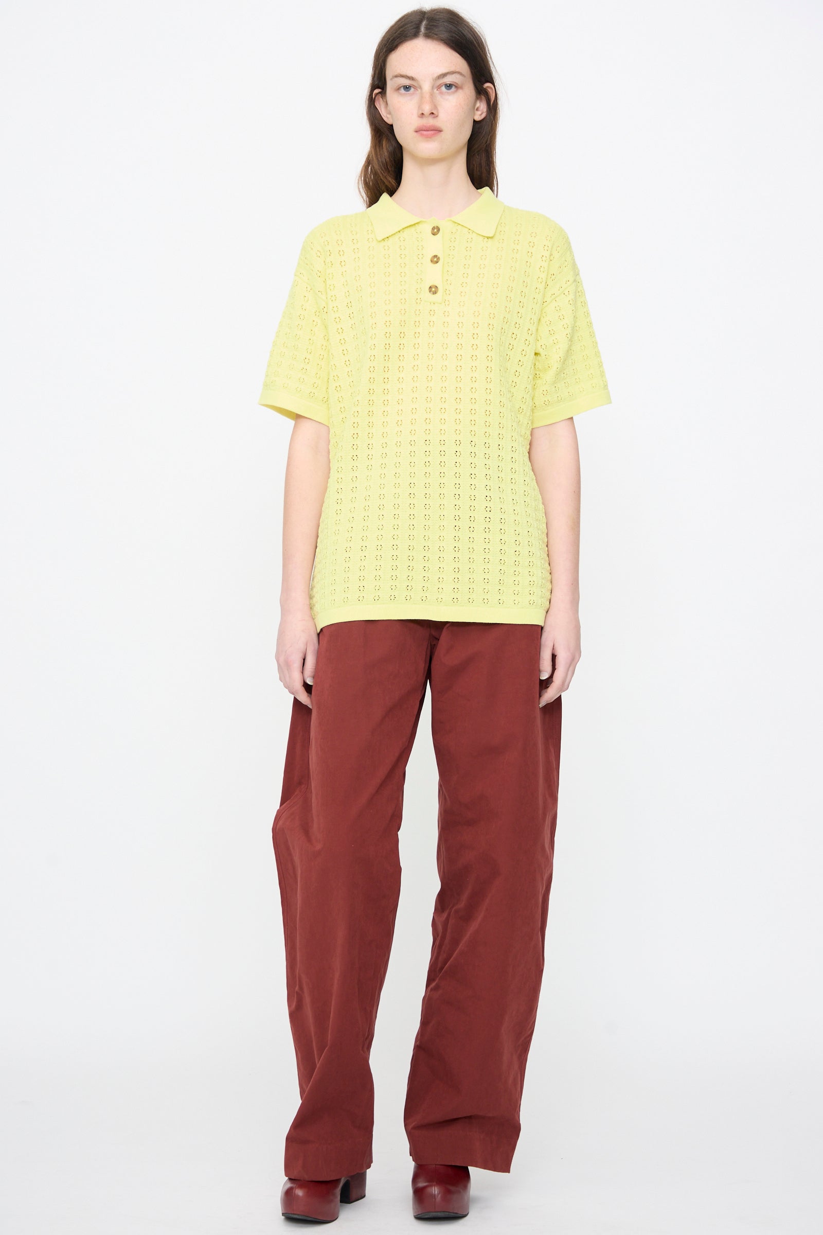 A person stands facing forward against a plain white background, wearing a yellow short-sleeve knit polo shirt, Rachel Comey Lyson Pant in Garnet, and matching maroon shoes.