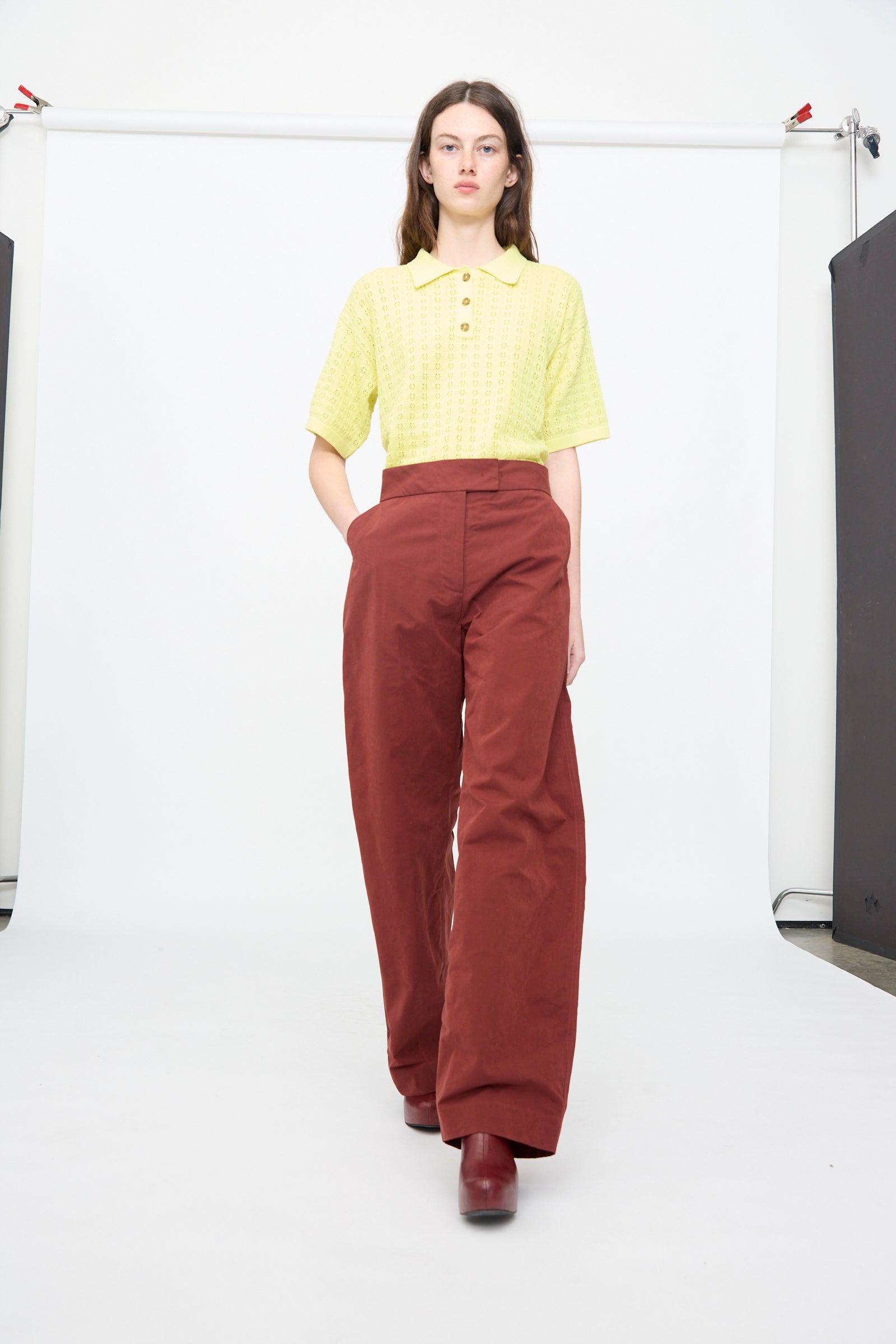 A person stands against a white backdrop wearing a light yellow short-sleeve collared shirt, Rachel Comey's Lyson Pant in Garnet, and maroon platform shoes.